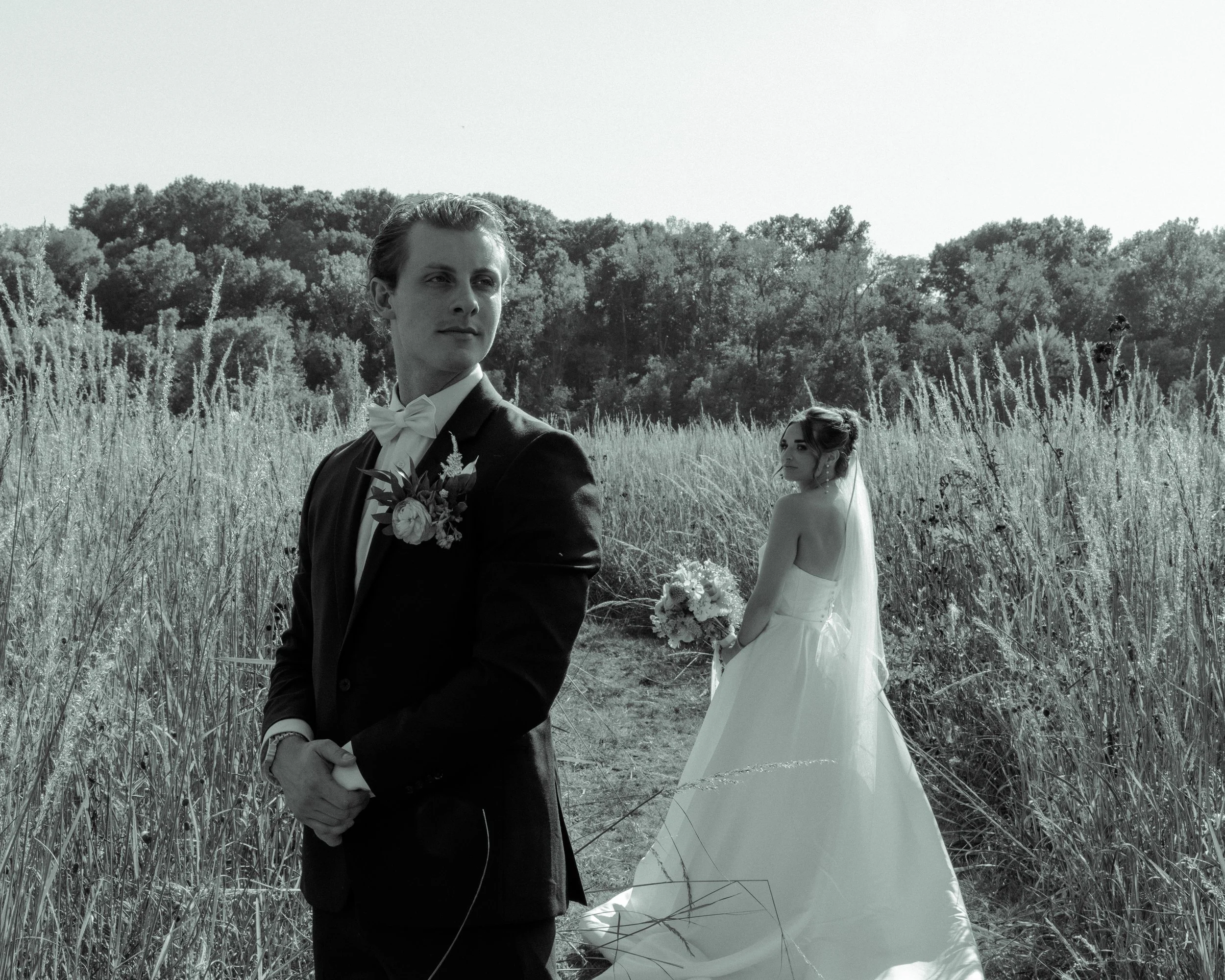 A groom in a tuxedo standing in a field with tall grass, looking to the side, with a bride in a white wedding gown holding a bouquet behind him, in a rural outdoor setting with trees in the background.