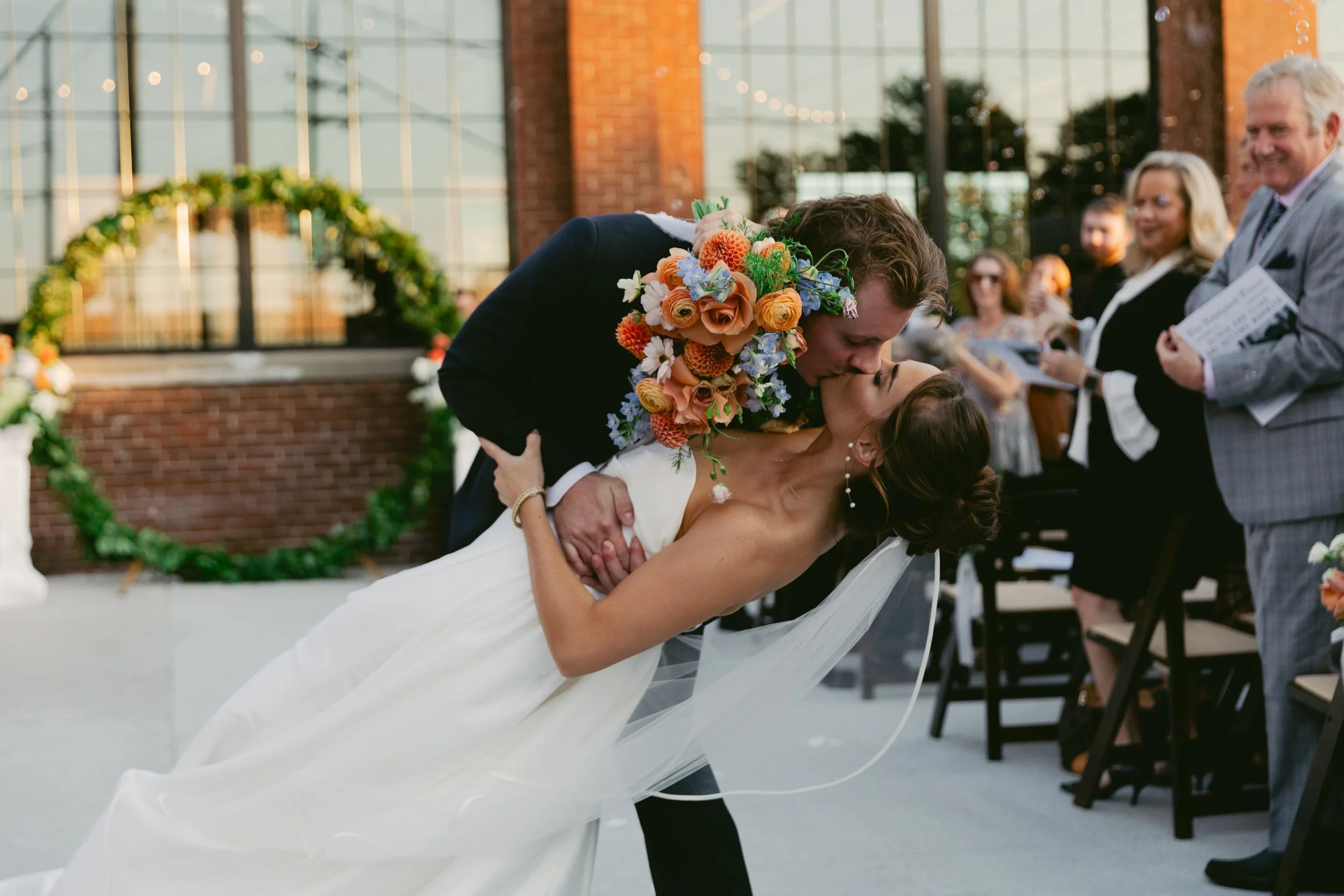 A newlywed couple sharing a kiss during their wedding ceremony, with guests smiling and observing in the background outside a brick building decorated with floral arrangements. St. Louis Documentary-style wedding photography on 35mm film, 120 film, a