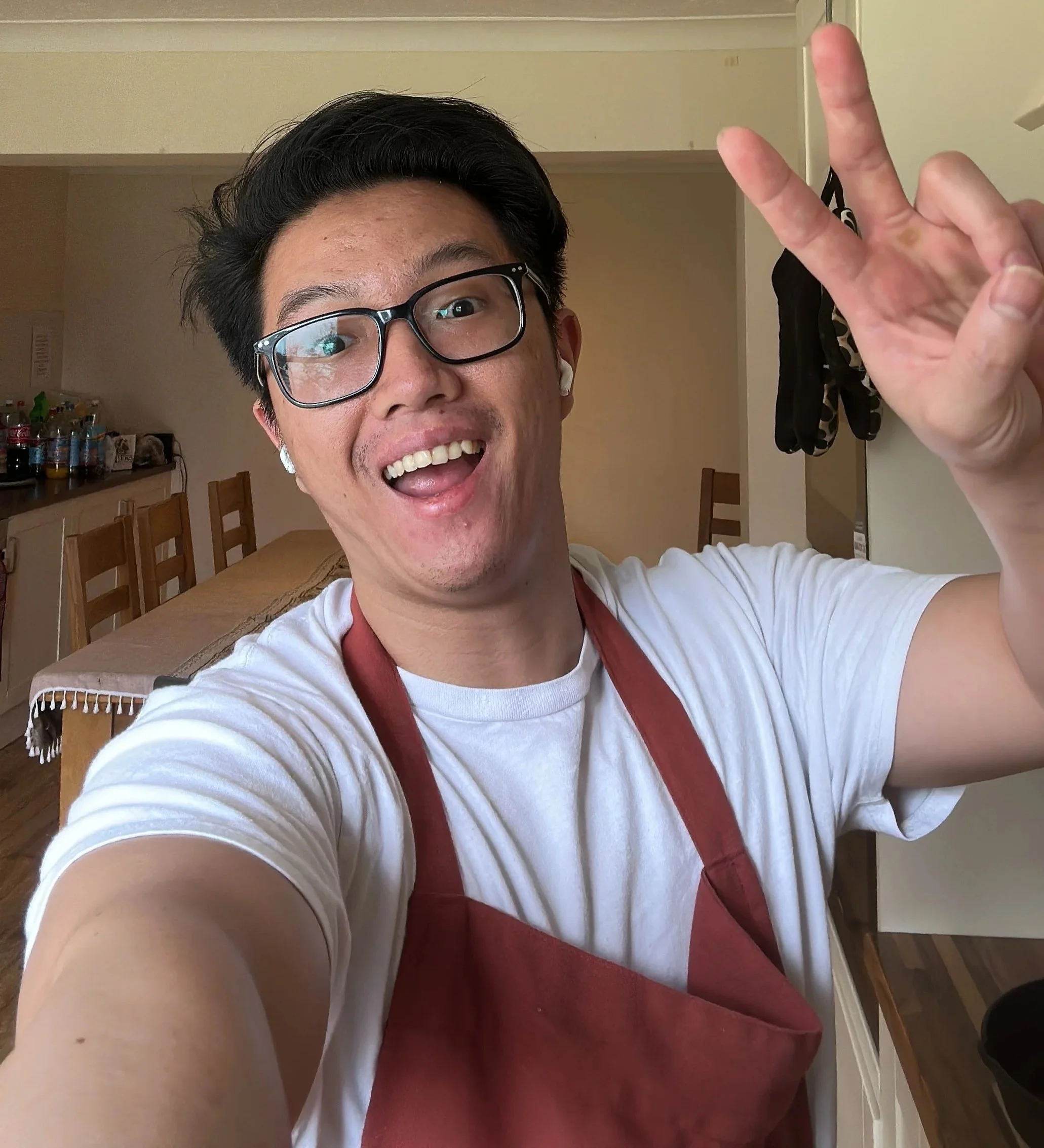 Rhedge with glasses and AirPods taking a selfie in a kitchen, making a peace sign with his right hand, and smiling.