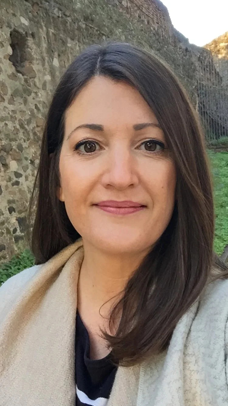 A woman with shoulder-length brown hair and brown eyes smiling outdoors with a stone wall and greenery in the background.
