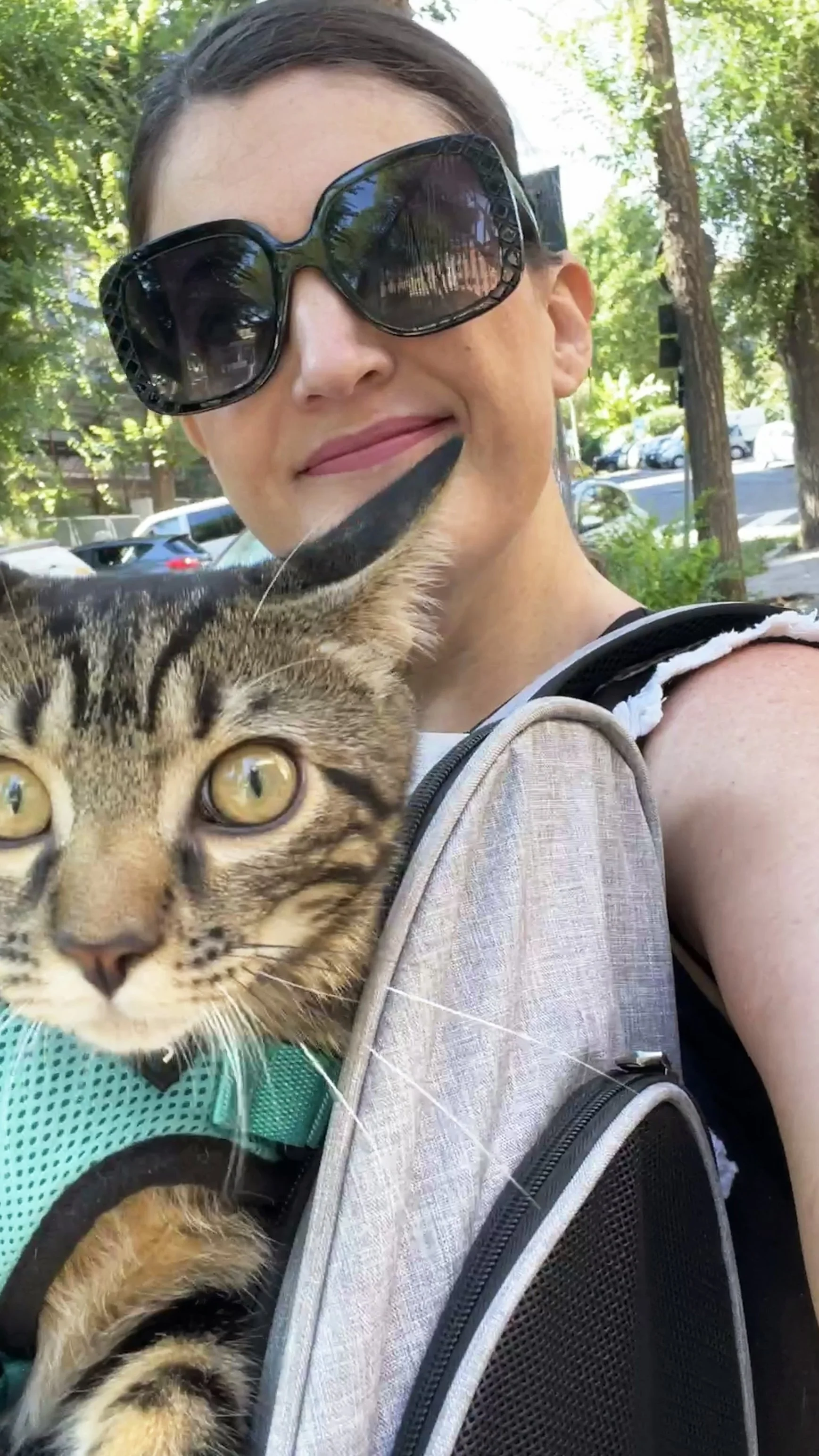 A woman with dark sunglasses smiling, holding a tabby cat in a harness on her shoulder outdoors with trees and parked cars in the background.
