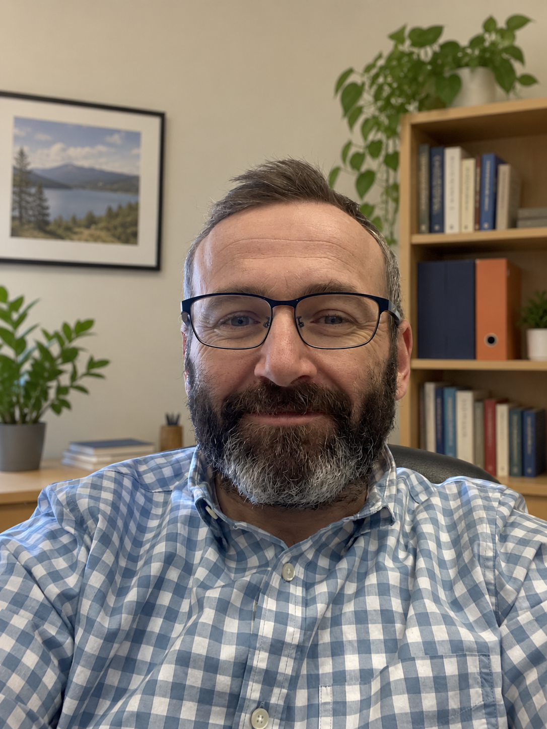 A man with glasses and a beard smiling for a selfie in an office with bookshelf, plants, and framed landscape photograph.