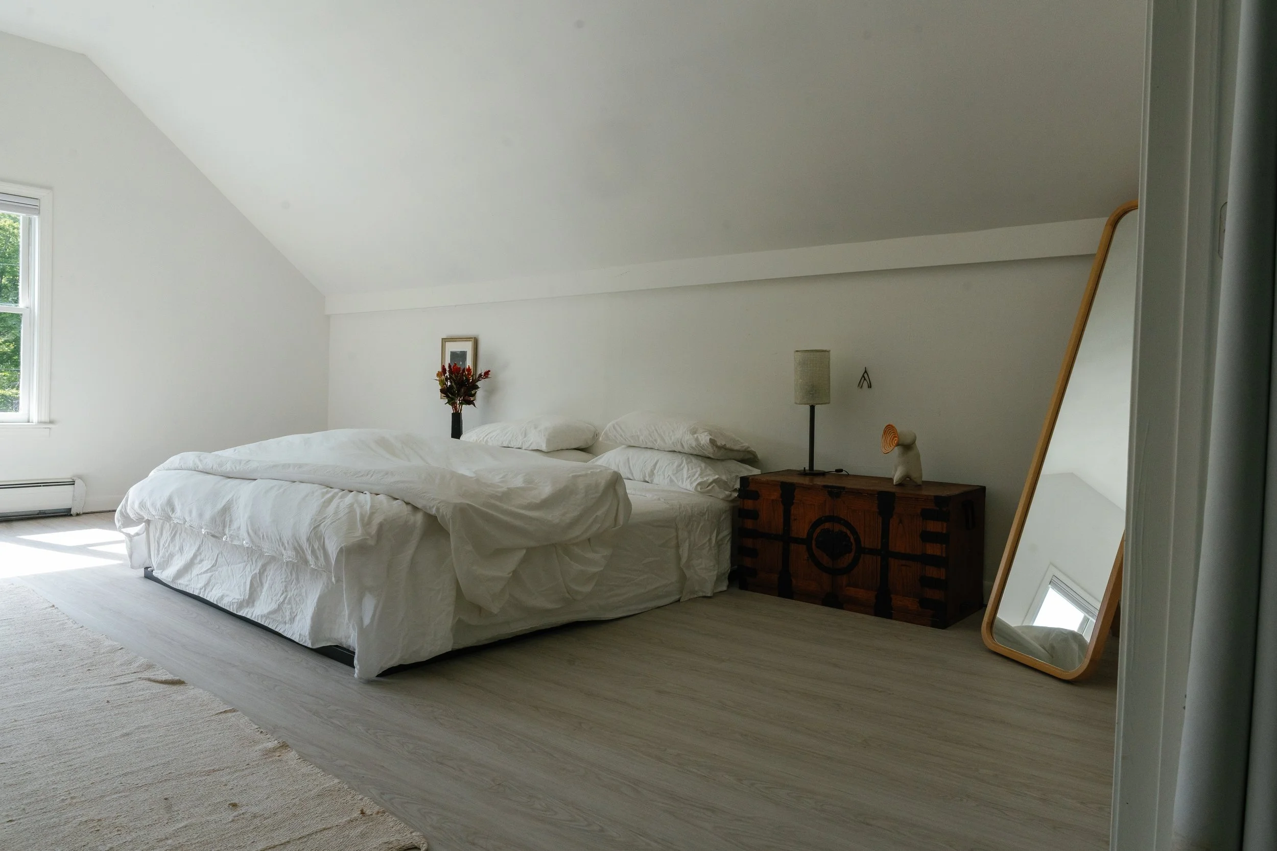 A cozy bedroom with white walls, a bed with white bedding, a vase with red flowers, a wooden nightstand, a tall floor lamp, and a large mirror leaning against the wall. A window with greenery outside brightens the room.