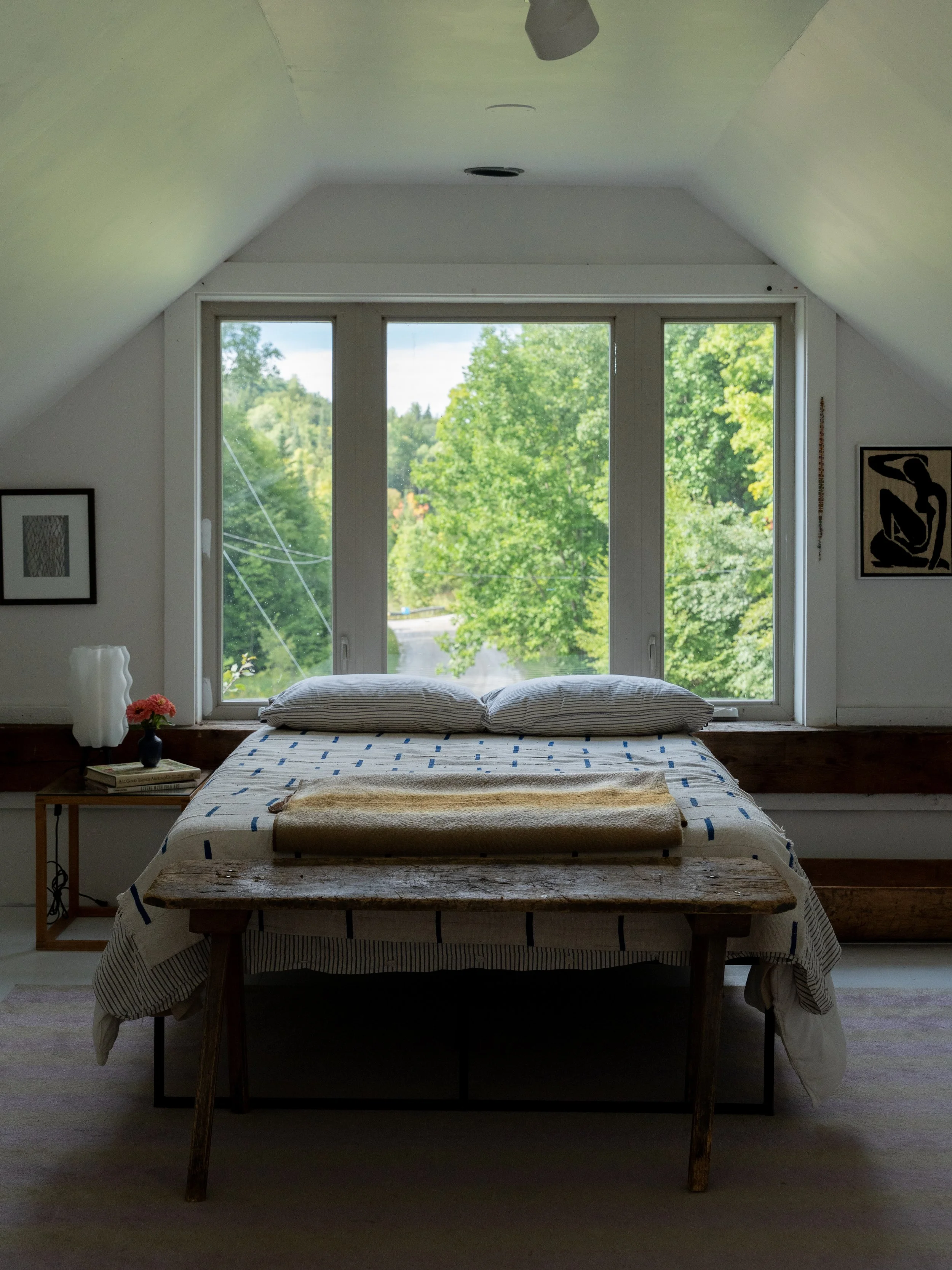 A bedroom with a large window showing green trees outside, unmade bed with striped bedding, pillows, and a blanket, wooden furniture, and framed artwork on the walls.