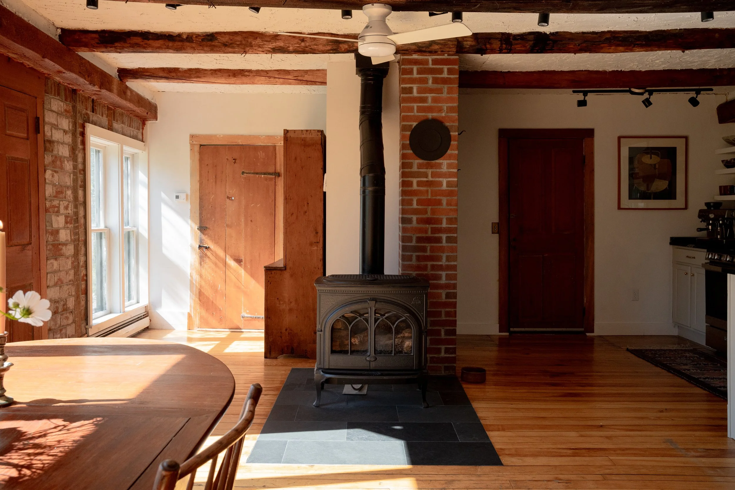 Interior of a rustic living room with exposed wooden beams on the ceiling, a wood stove, a brick chimney, wooden doors, and natural light coming through windows.