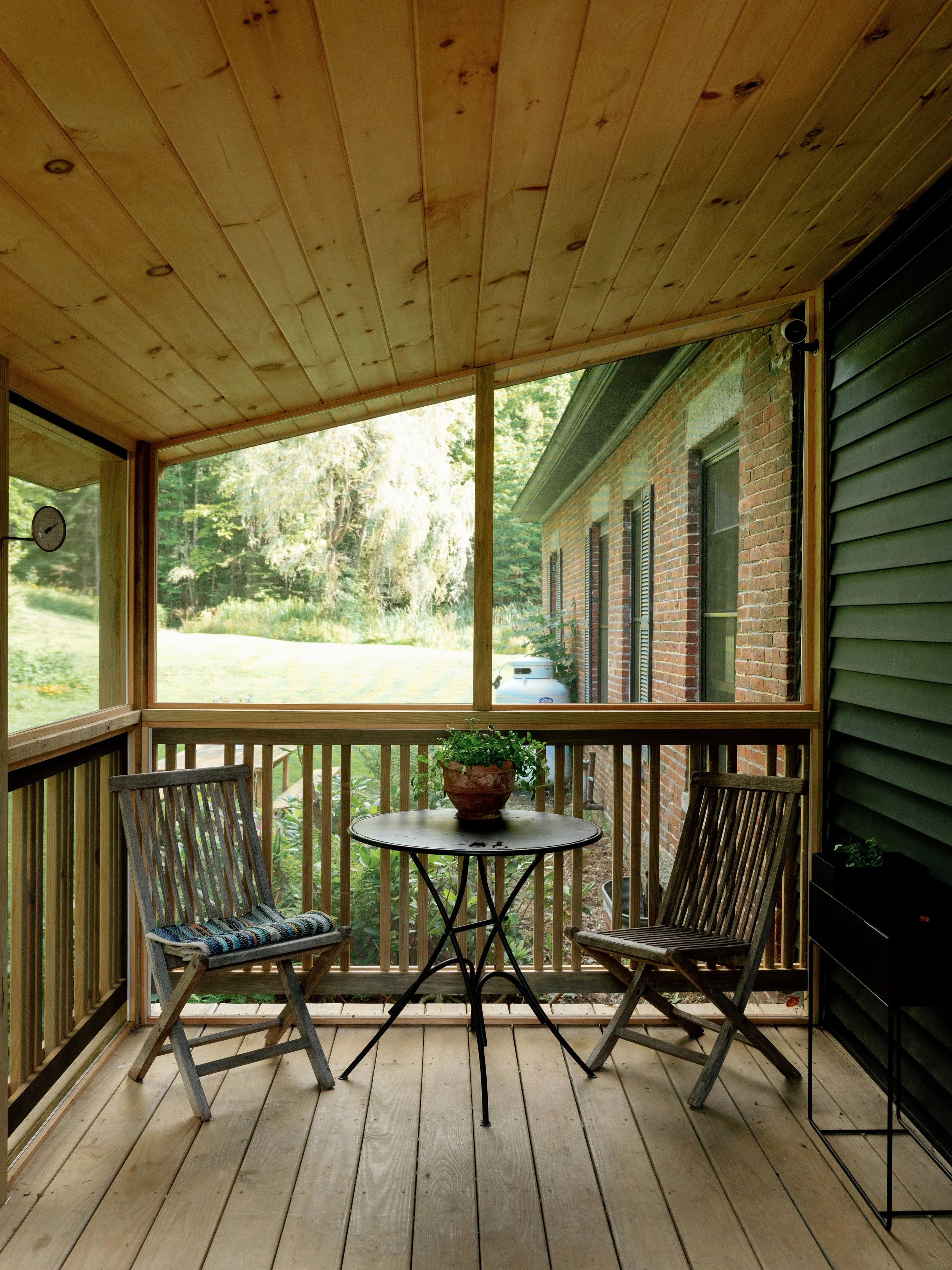 A cozy screened-in porch with wooden flooring, a wooden ceiling, and a view of lush greenery outside. There are two wooden chairs with cushions, a small round table with a potted plant, and a side cabinet on the right. The porch is attached to a bric