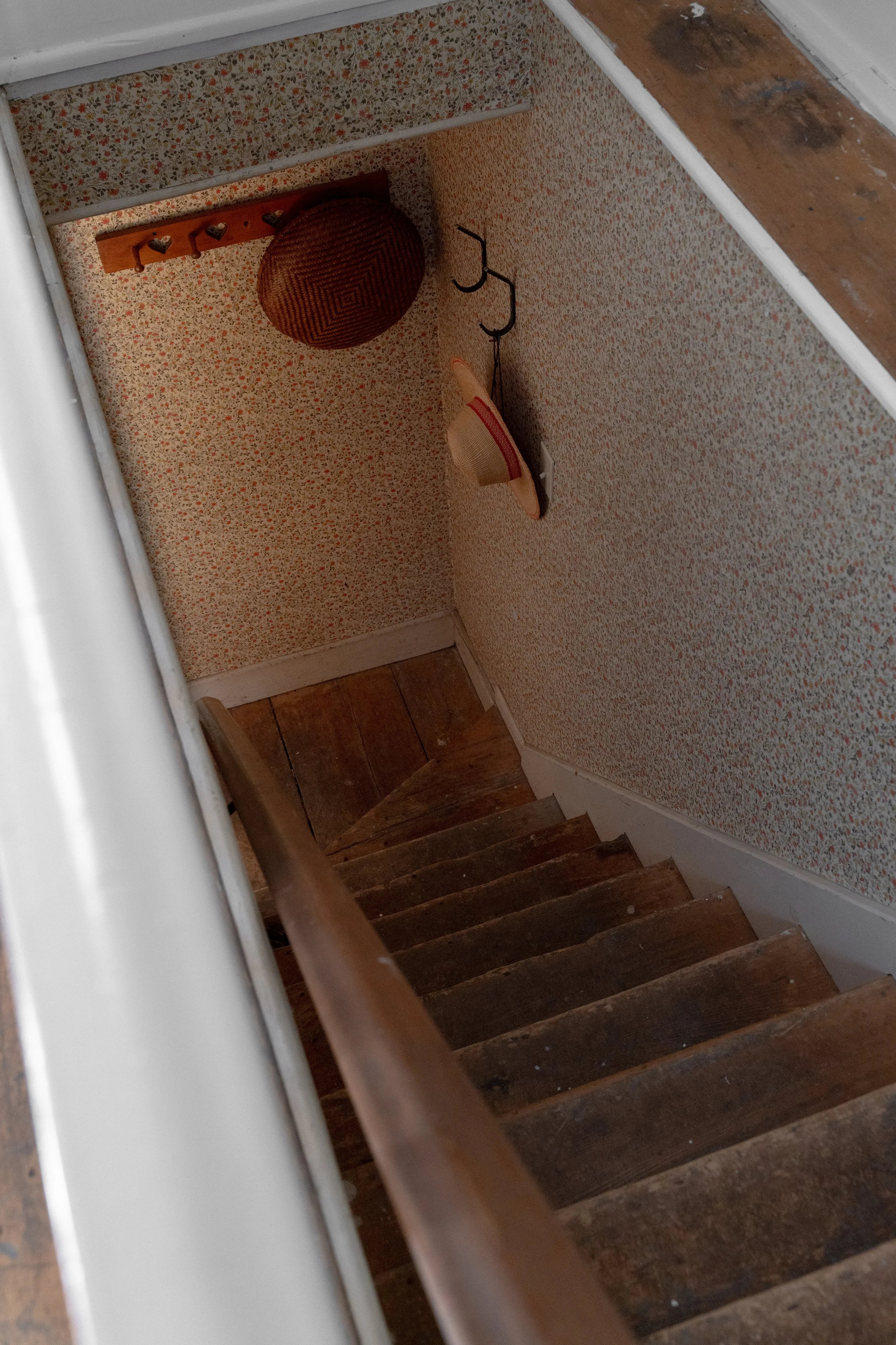 View of a wooden staircase with dark stained steps, beige floral wallpaper on the wall, a wicker basket, a straw hat, hooks, and a small black hook-like object.