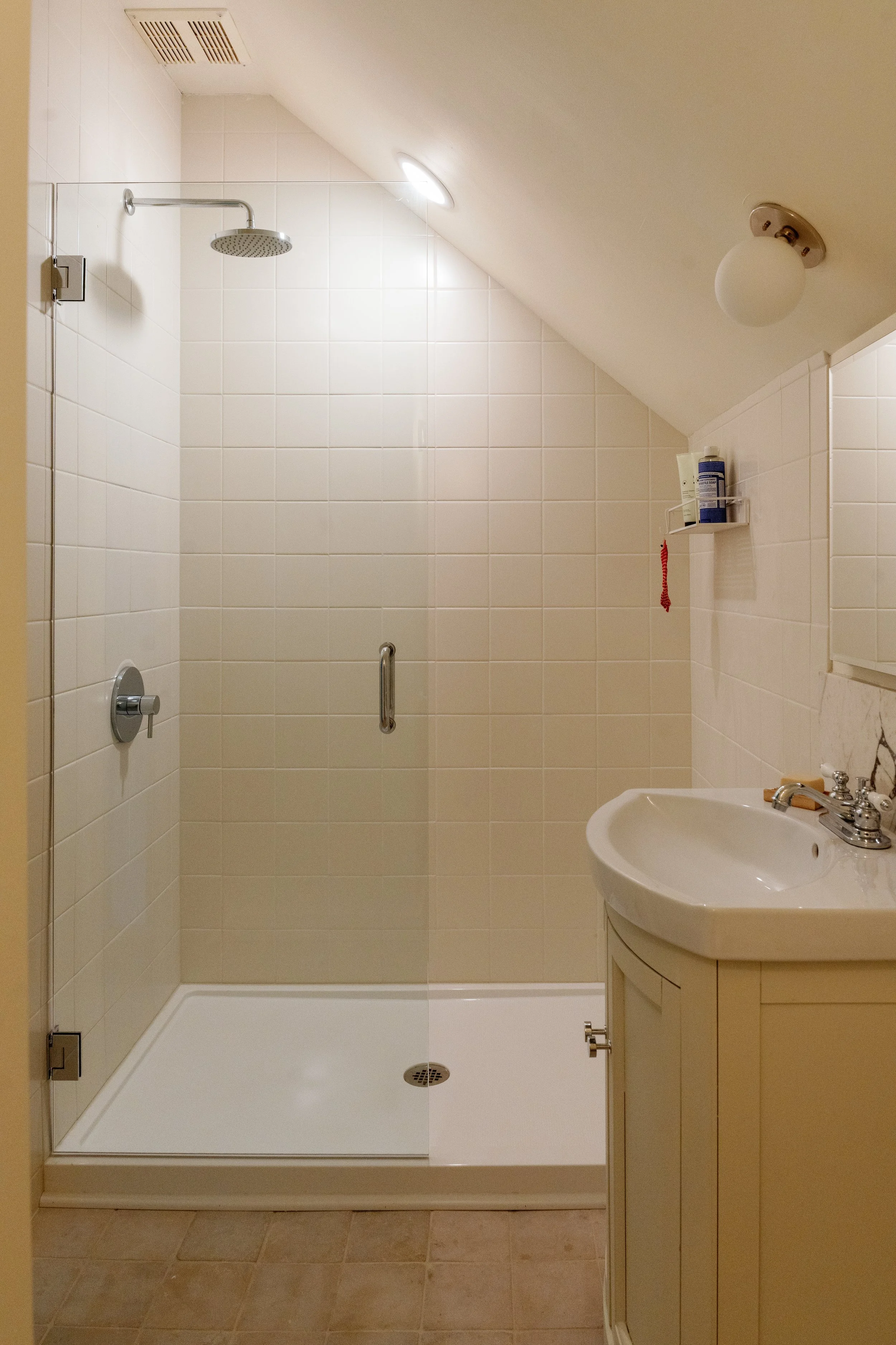 Bathroom with a walk-in shower, a small sink with a cabinet, and a mirror. The shower has a glass door, a rain showerhead, and a wall-mounted shower control. There is a shelf in the shower holding bottles. The ceiling is sloped with a light fixture, 