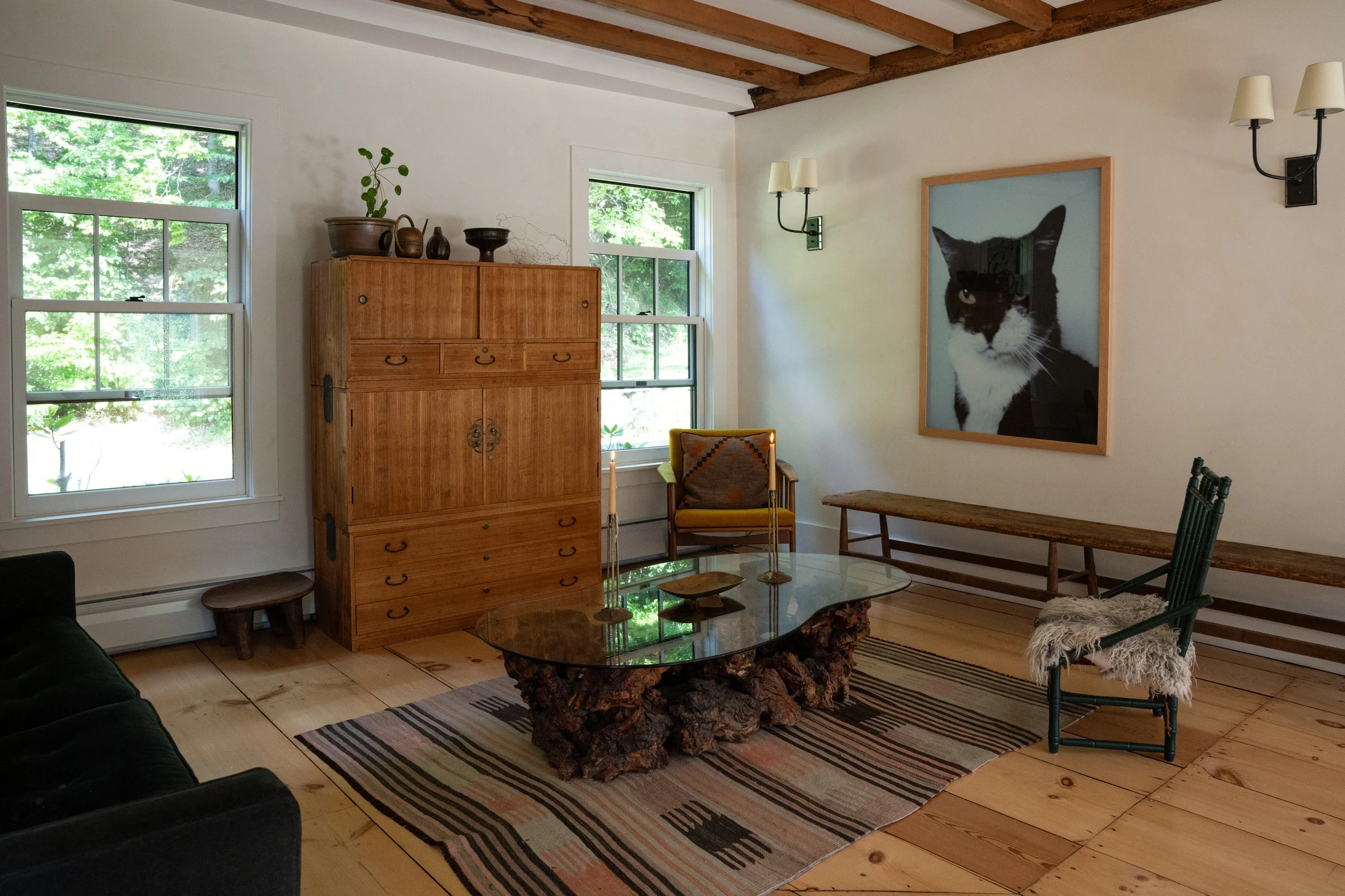 A cozy living room with two large windows showing green trees outside. The room features wooden beams on the ceiling, a wooden cabinet with pottery on top, a glass coffee table with a textured base, a yellow armchair, a black couch, a large framed ph
