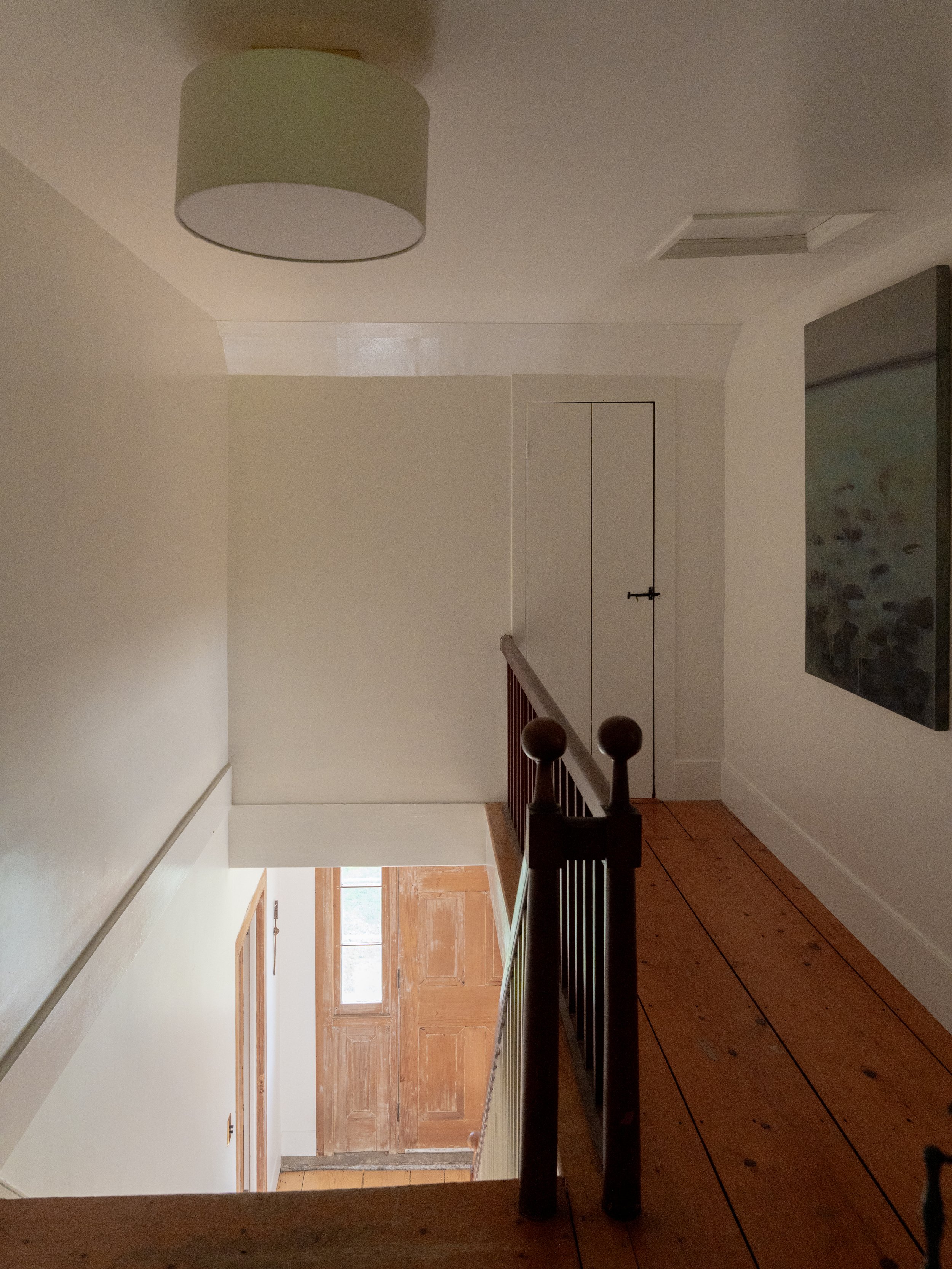 Interior view of a hallway with a wooden staircase railing, a closed wooden door, and a painting on the wall.