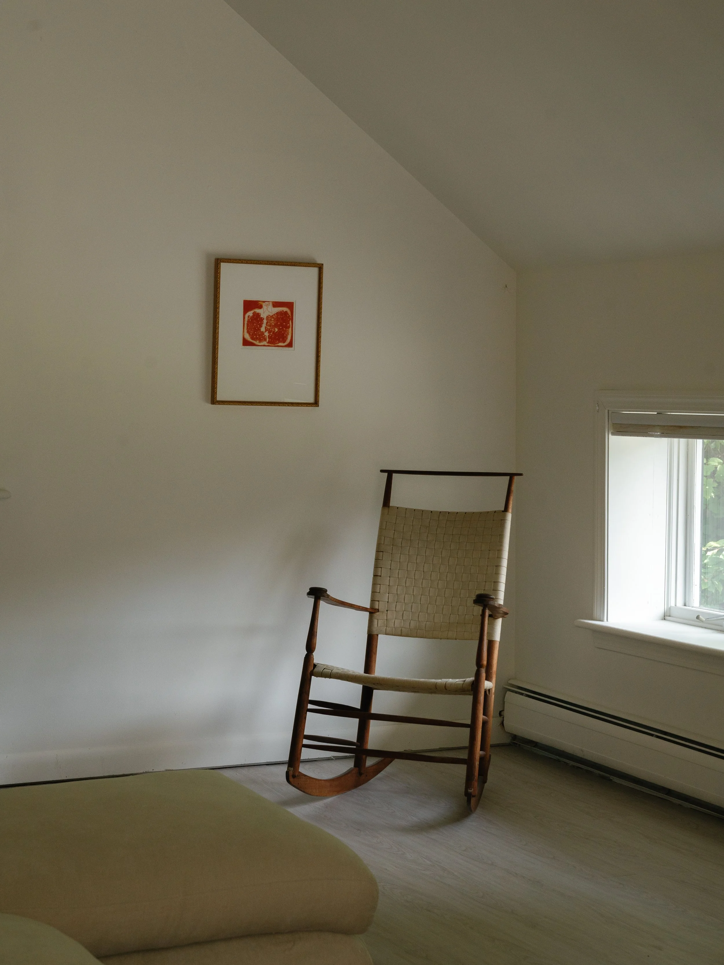 A wooden rocking chair on a wooden floor near a window in a plain room with white walls and a framed picture of a pomegranate on the wall.