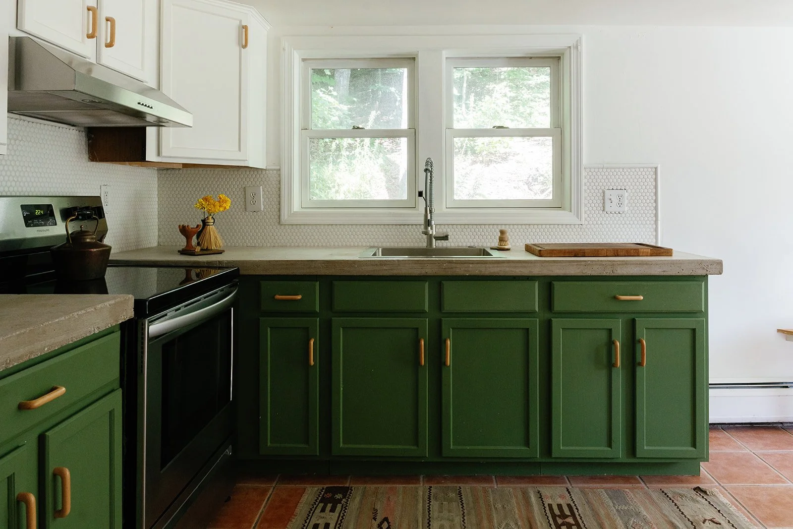 Kitchen with green lower cabinets, white upper cabinets, a window above the sink, a black stove, and a beige countertop with a cutting board. There are yellow flowers on the counter and a teapot near the stove.