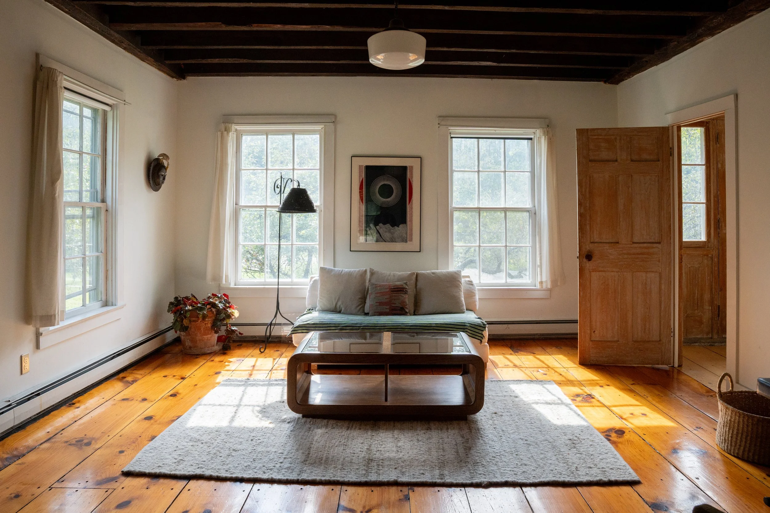 A cozy living room with white walls, large windows letting in natural light, a sofa with striped cushions, a coffee table, a potted plant, and a framed artwork on the wall. Wooden beams on the ceiling and hardwood flooring add warmth to the space.