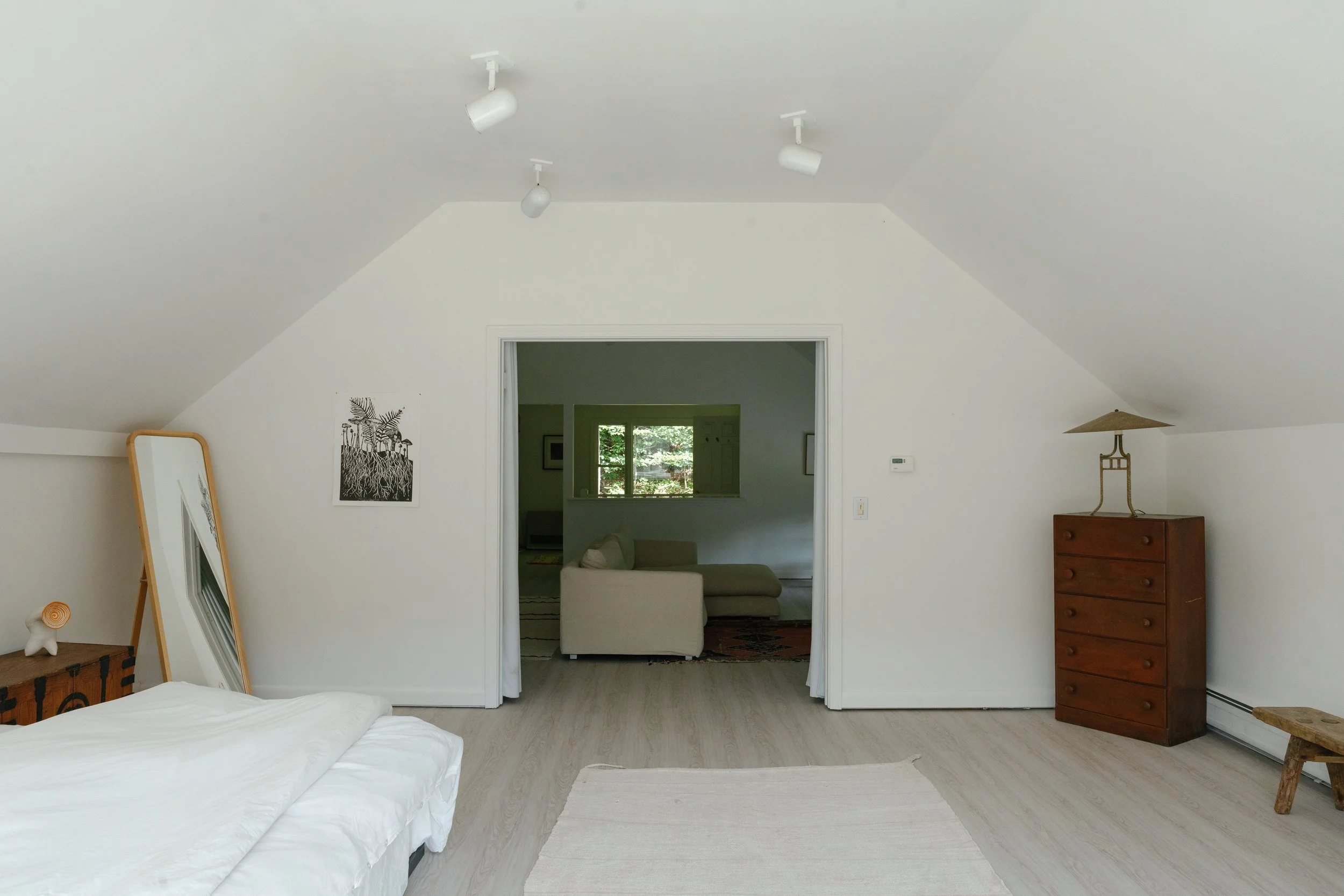 Interior of a bedroom with a white sloped ceiling, a bed with white bedding in the foreground, a mirror leaning against the wall, a small table with a lamp to the right, and a doorway leading to a living room with a beige sofa and window in the backg