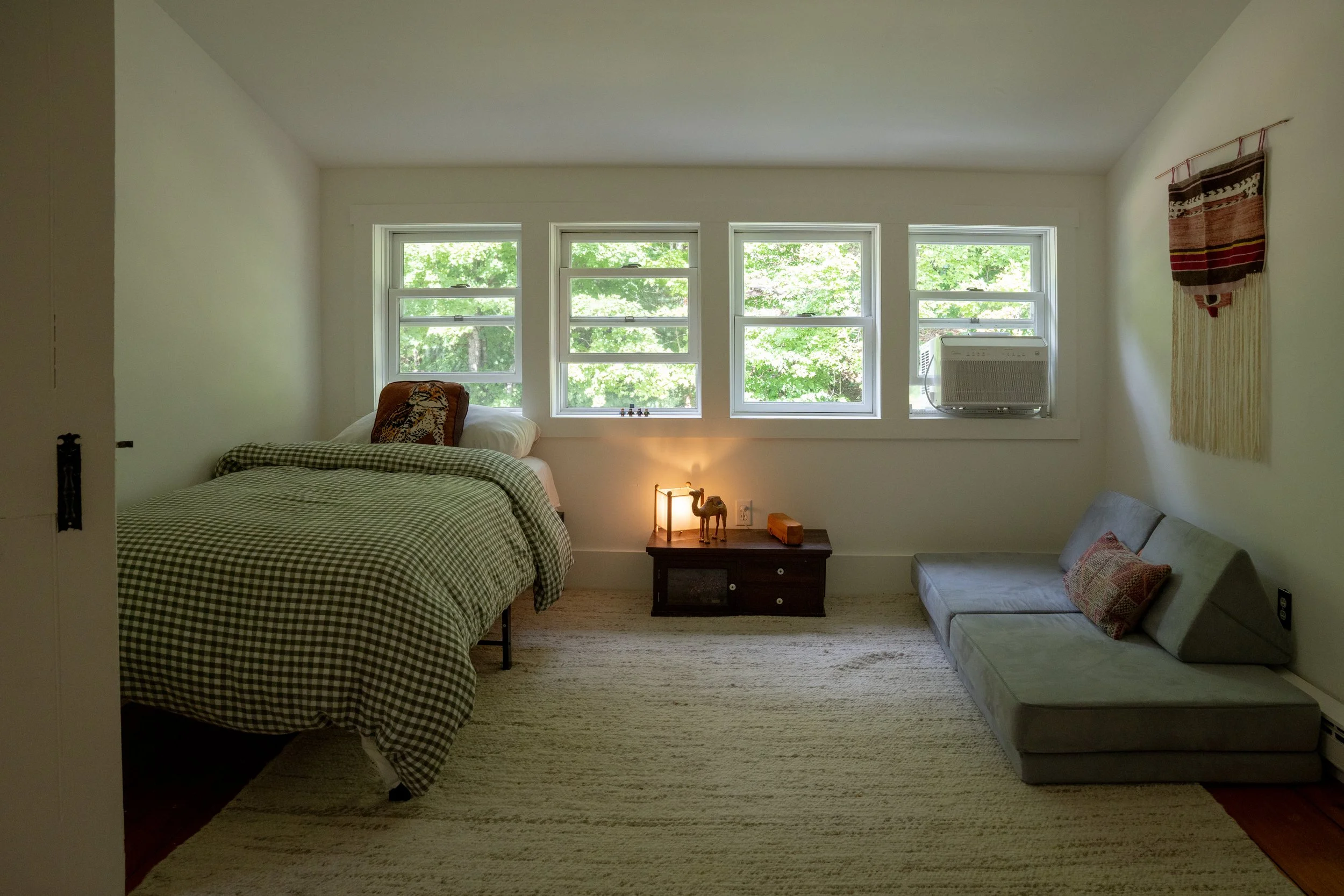 A cozy bedroom with a bed featuring a black and white checkered bedspread, a pillow with a tiger print, and a matching throw pillow. The room has a set of four windows letting in natural light and offering a view of green trees outside. On the right,