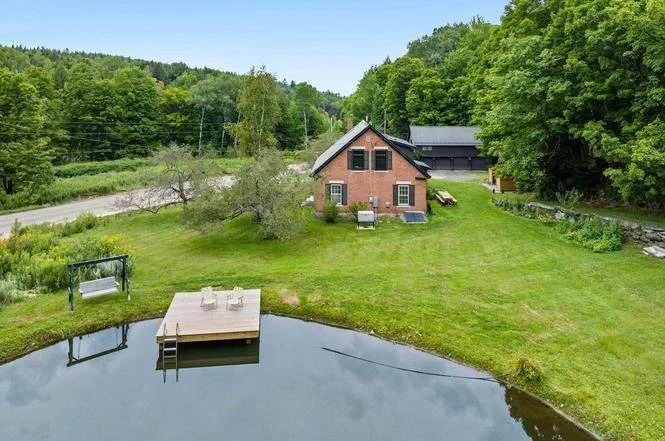 A house with a brick exterior and a black roof, surrounded by a green lawn and trees. There is a small pond with a dock and two chairs, and a swing set nearby. The setting appears to be in a rural area with a road and forest in the background.