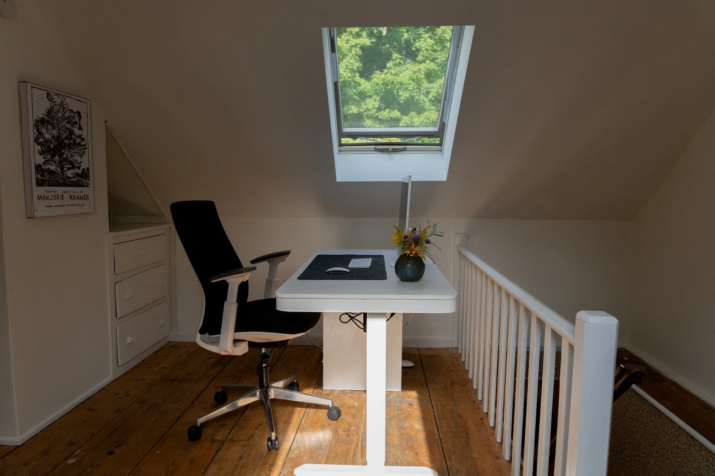 A white desk with a computer, mouse, and a vase of flowers, placed under a skylight window in an attic room with wooden floors and a white railing.