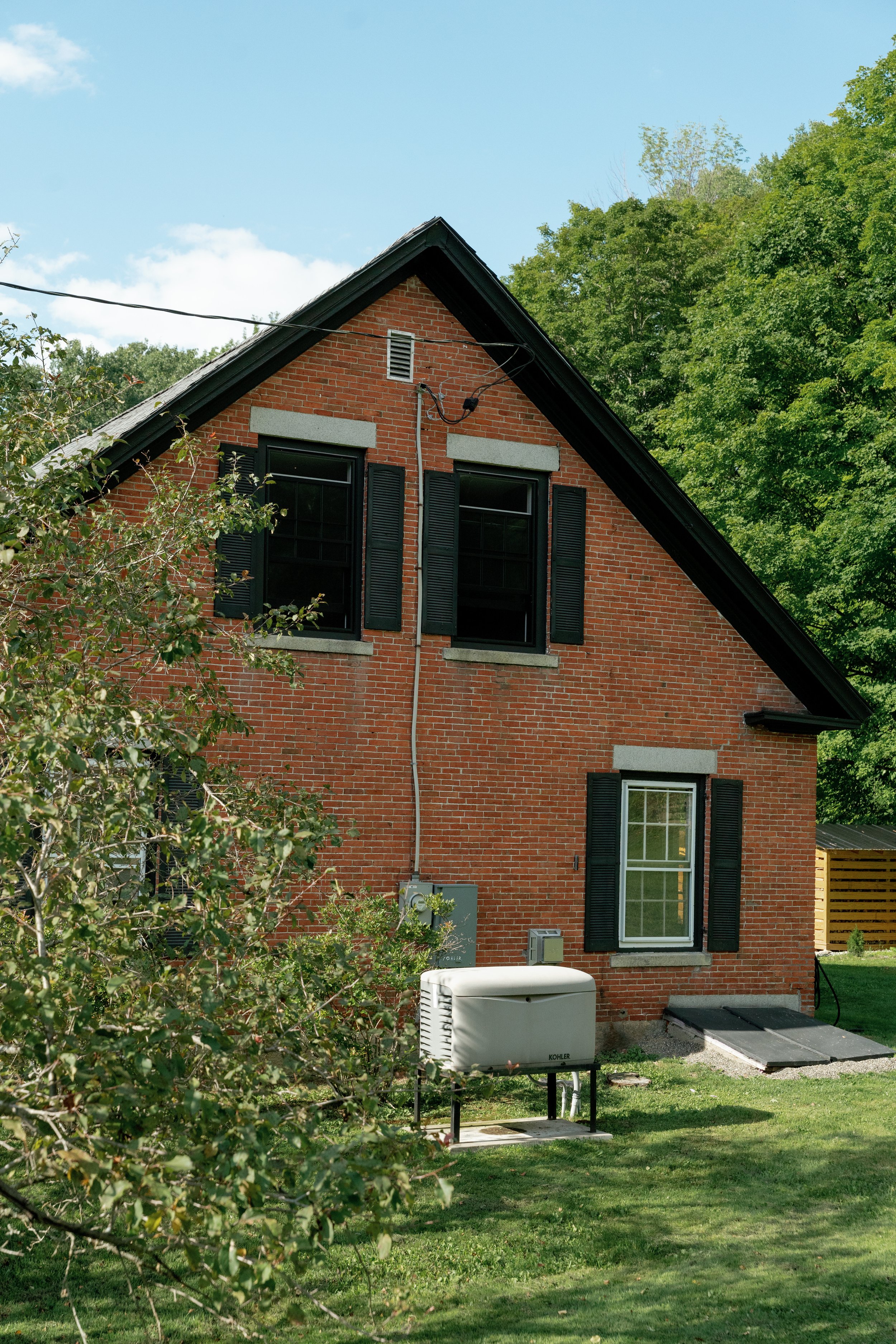 A red brick house with black shutters and a steep roof, surrounded by green trees and grass. There are utility boxes and a white HVAC unit outside, with a cloudy sky overhead.