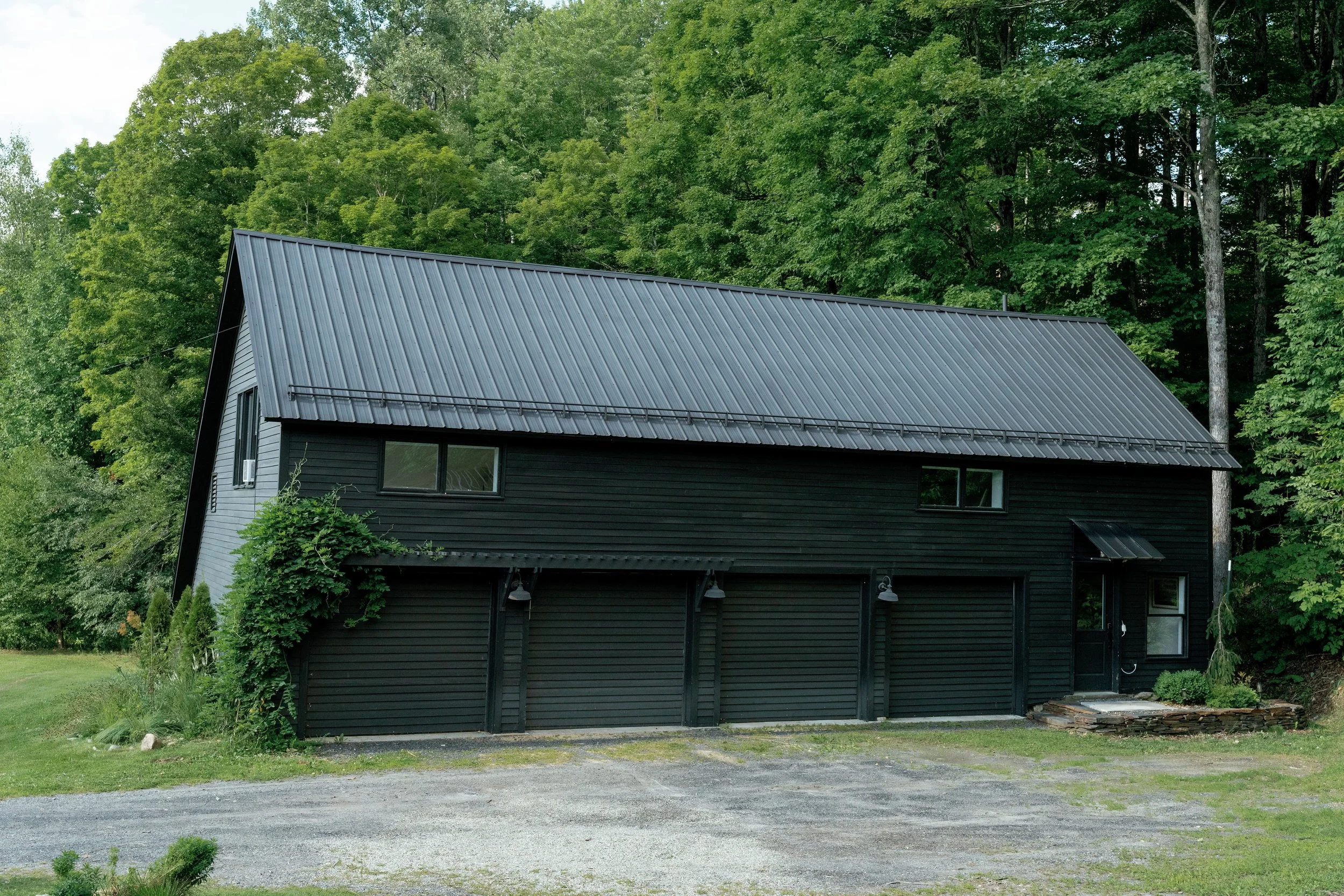 A modern house with black exterior walls and a steep metal roof, surrounded by lush green trees.