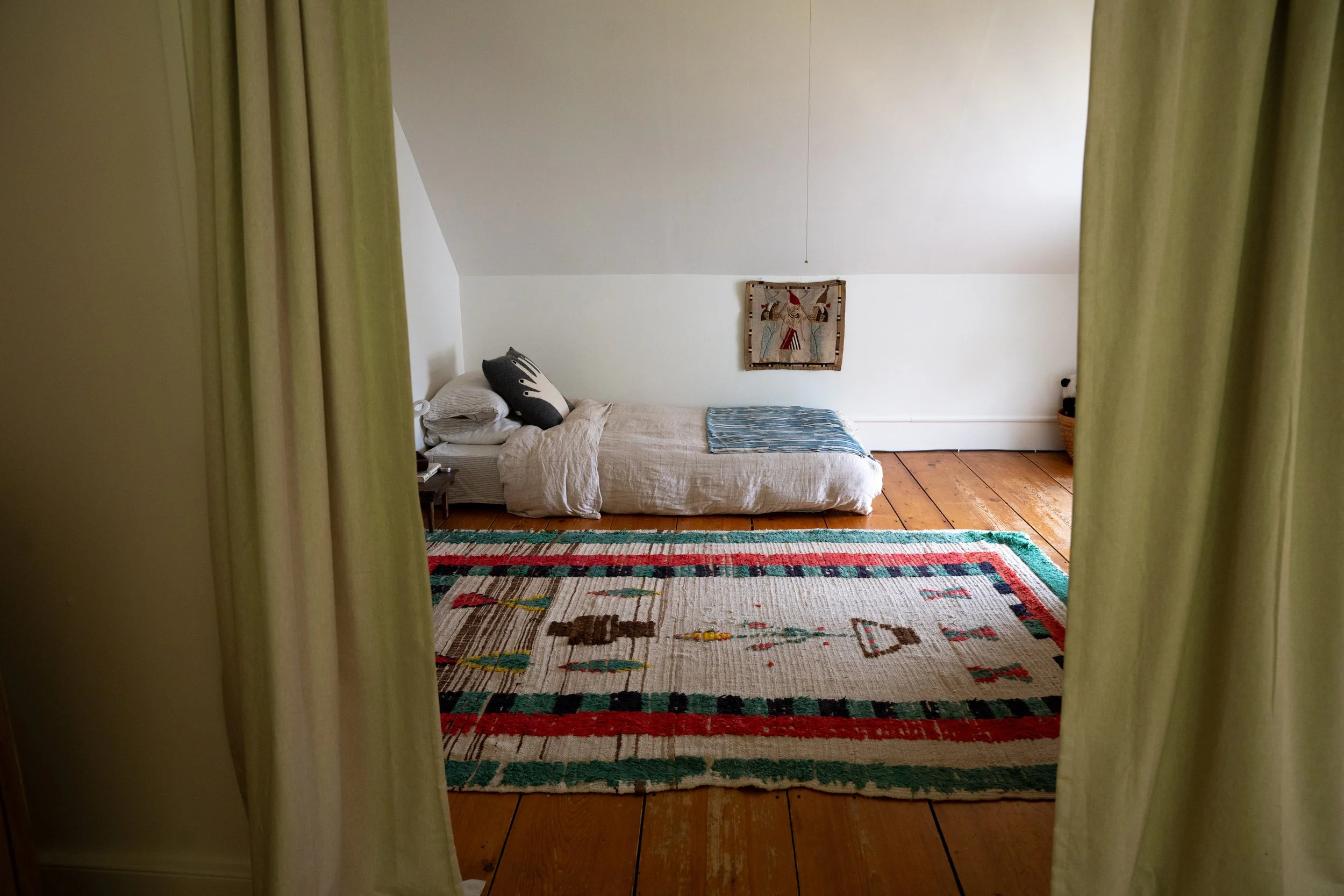 View of a cozy bedroom seen through green curtains, with a bed against a white wall, a colorful patterned rug on wooden floors, and a small piece of artwork hanging above the bed.