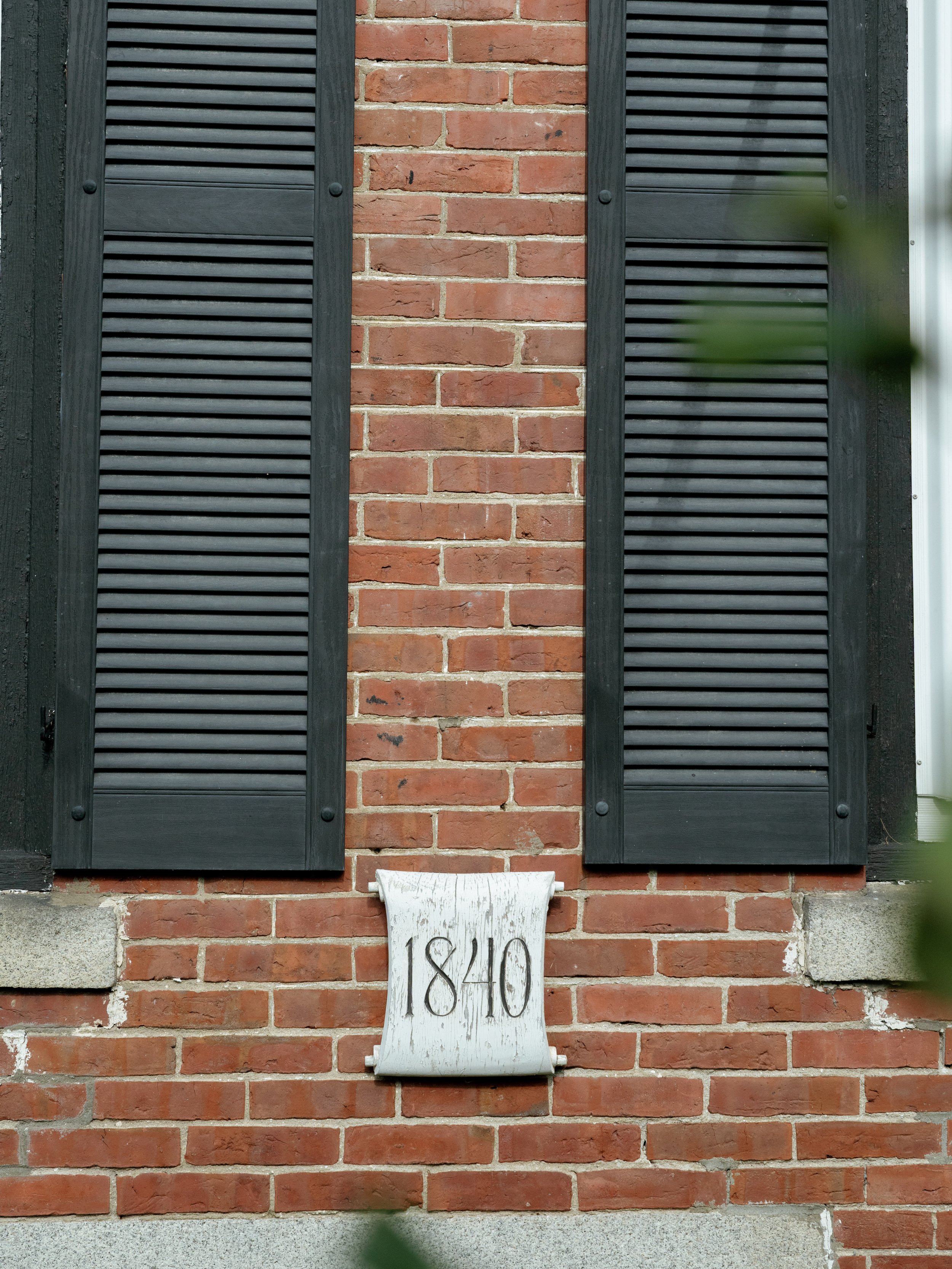 A brick house wall with black shutters on either side of a white wooden plaque that has the year '1840' written on it.