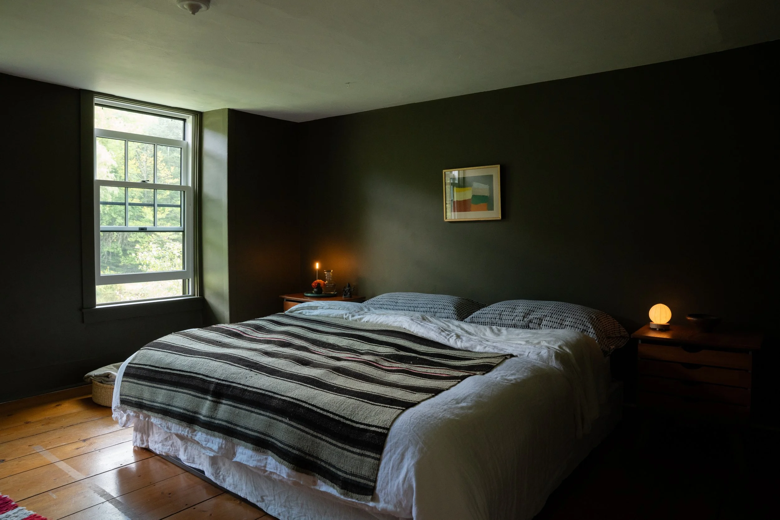 A cozy bedroom with a large bed covered with a striped black, white, and gray blanket, a window revealing green trees outside, a nightstand with a small lamp, framed artwork on the wall, and wooden flooring.