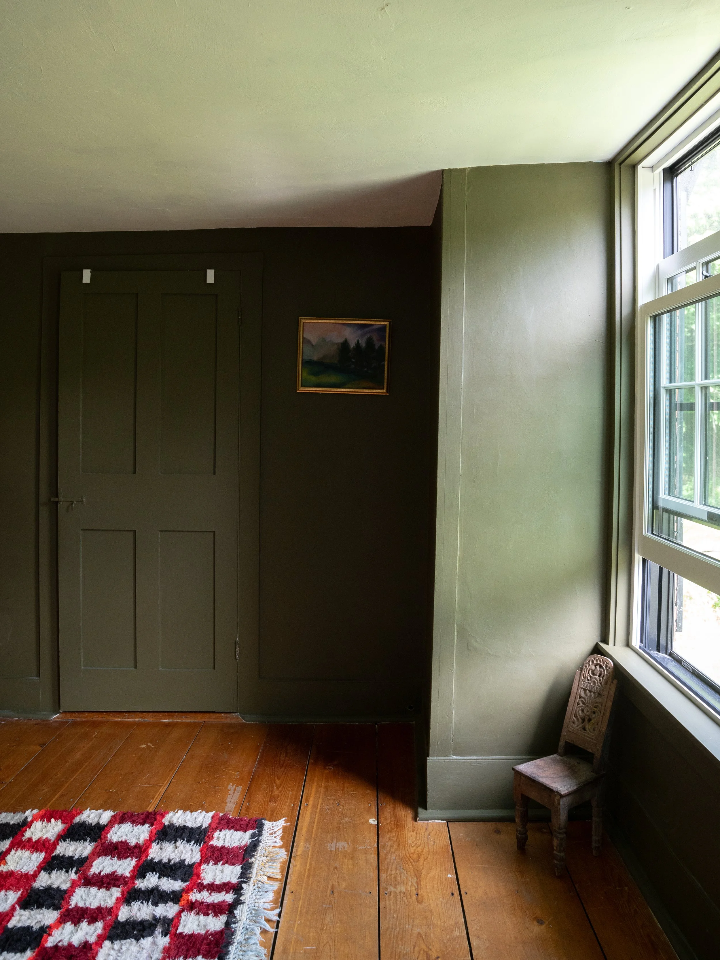 A room with wooden floor, green walls, a closed door, a small wooden chair, a colorful rug, and a window with a view of greenery outside.
