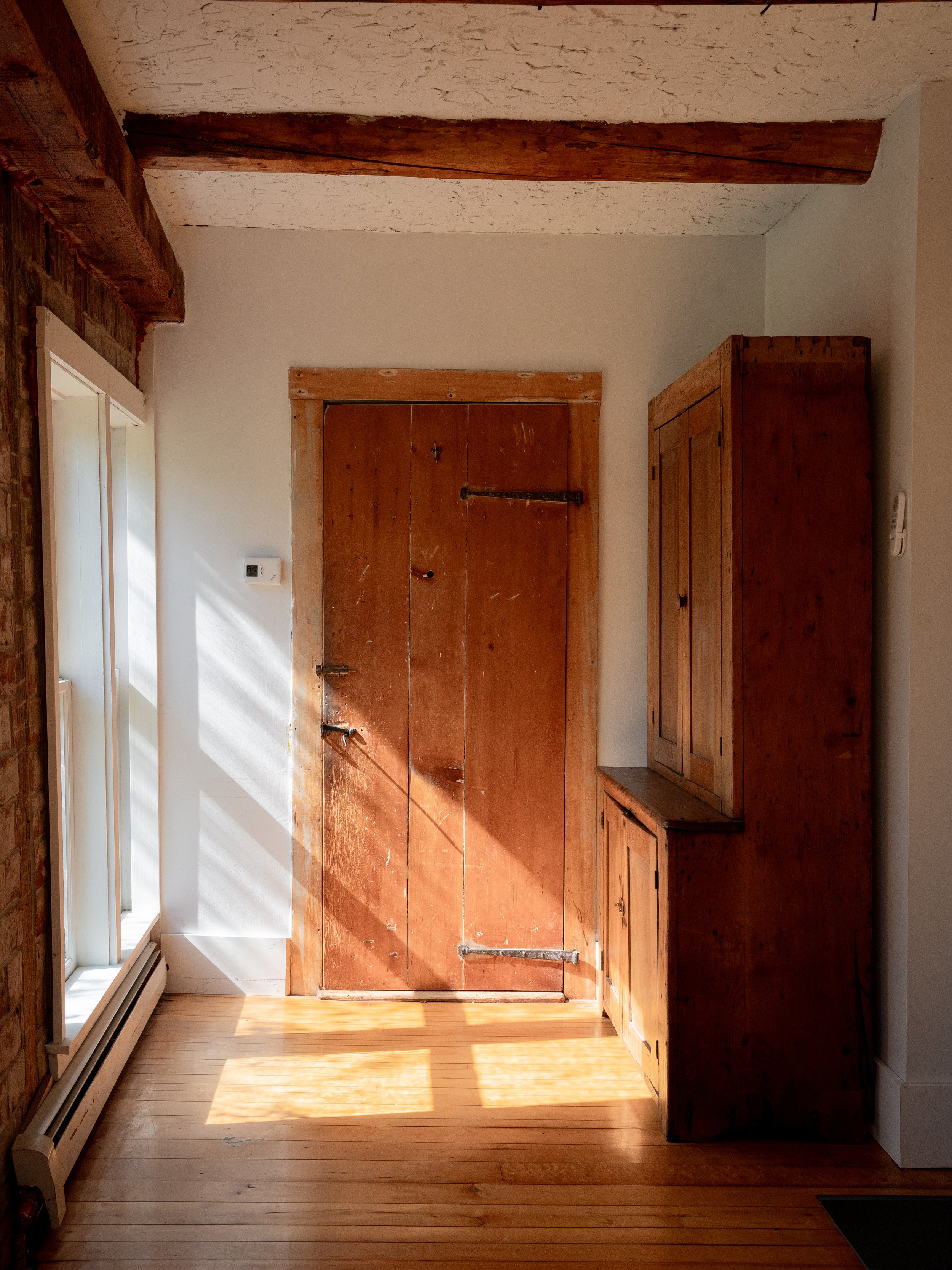 A room with wooden flooring and a wooden door, a wooden cabinet, and a window with white trim on the left side, natural light coming in.