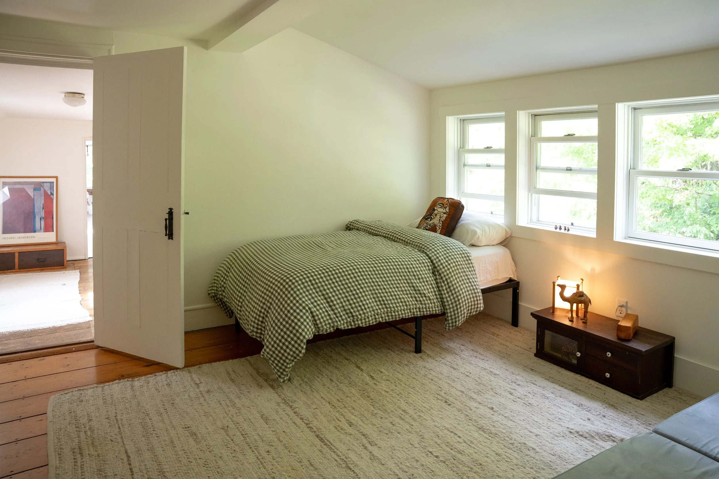 A bedroom with a bed covered in a checkered blanket, a small table with a lamp and dog figurine, large windows showing green trees outside, and a carpet on wooden floor.
