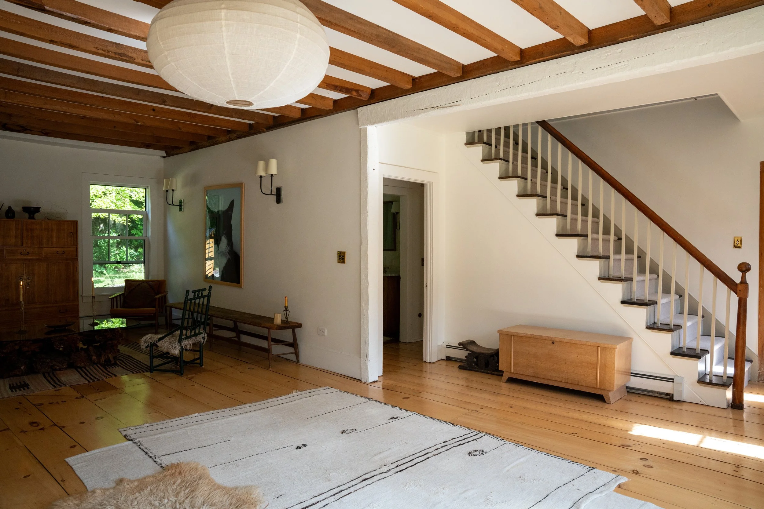 Interior of a living room with wooden floors and exposed wooden beams on the ceiling. There is a staircase with white risers and a wooden handrail. The room features a window with greenery outside, a wooden sideboard, wall sconces, and various chairs