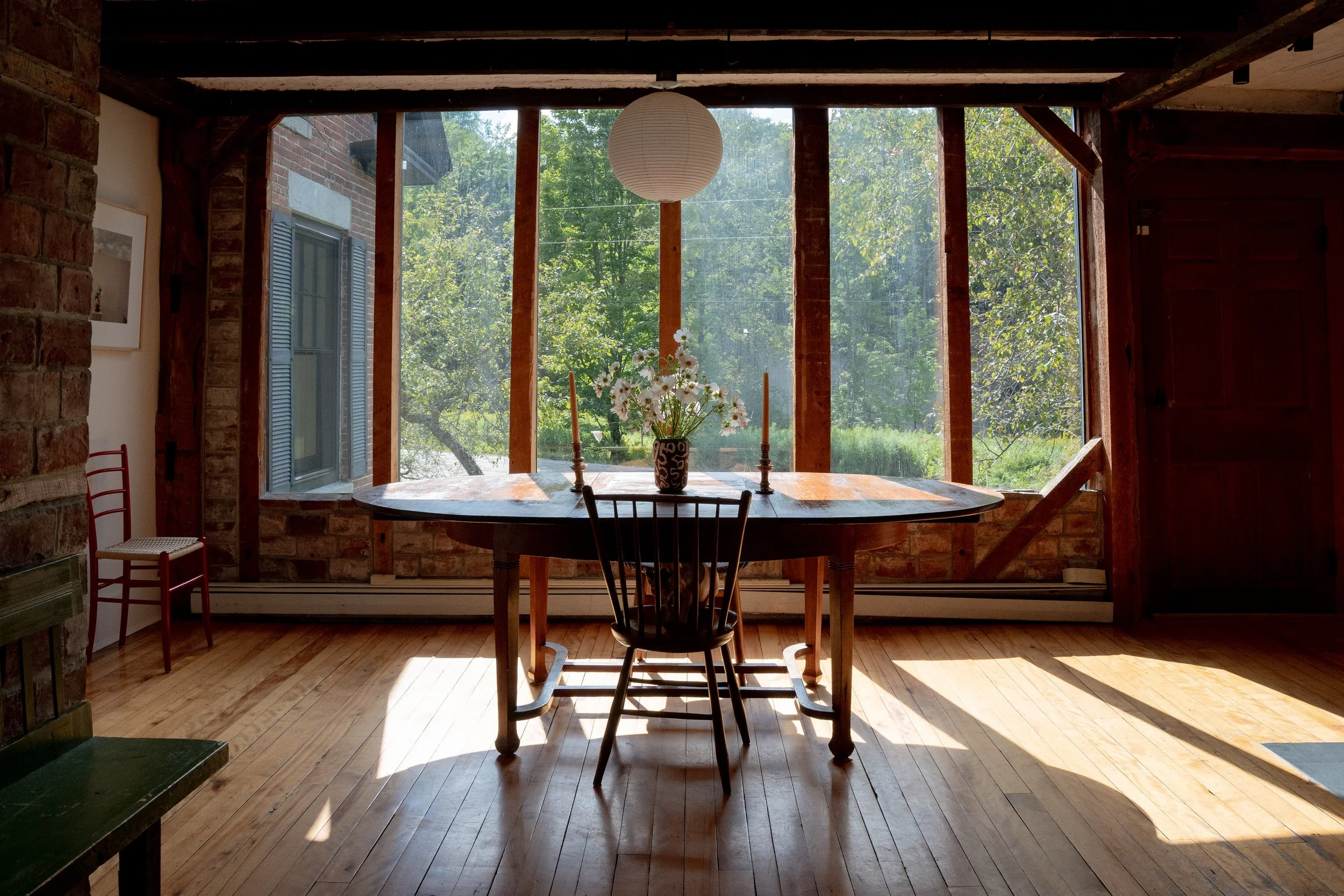 A rustic dining room with large windows overlooking green trees. There is a wooden table with a flower vase and two candlesticks, surrounded by chairs. The ceiling has exposed wooden beams and a paper lantern hanging at the center.