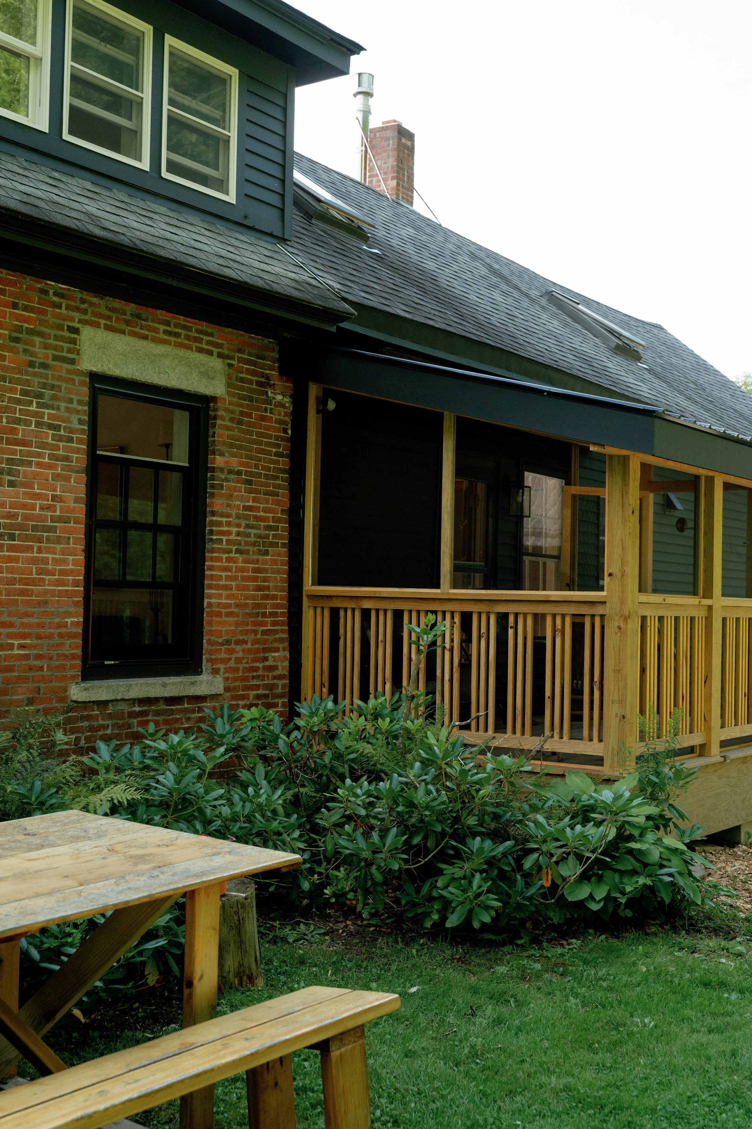 Partial view of a house with a brick exterior, black window frame, and a roof with dormer windows. There is a wooden deck or porch under construction with vertical wooden posts and railing. The yard has green bushes, grass, and garden plants.