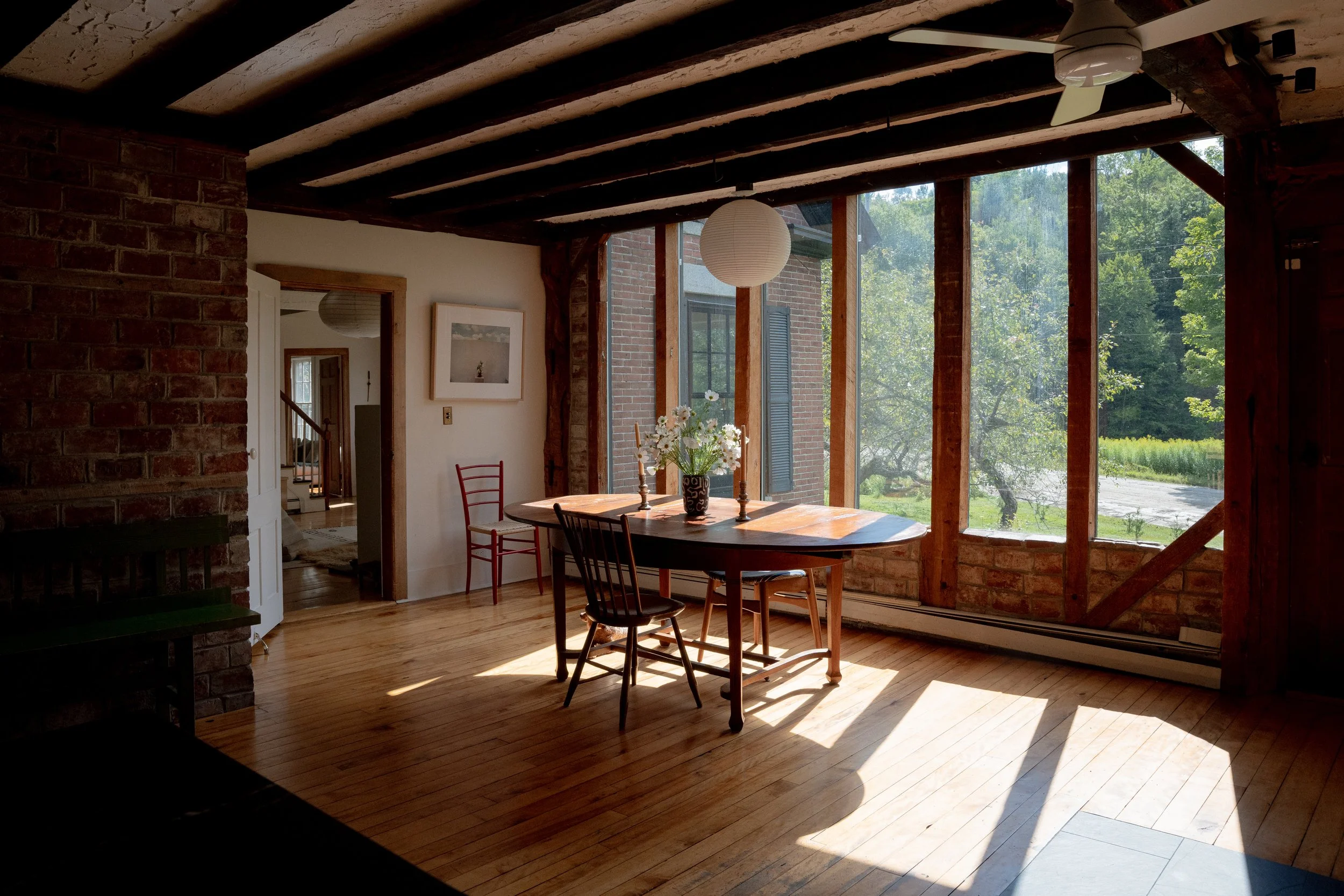 Interior of a rustic dining room with exposed brick and wooden beams, featuring a round wooden table with a floral centerpiece, surrounded by black and red chairs, and large windows with greenery outside.