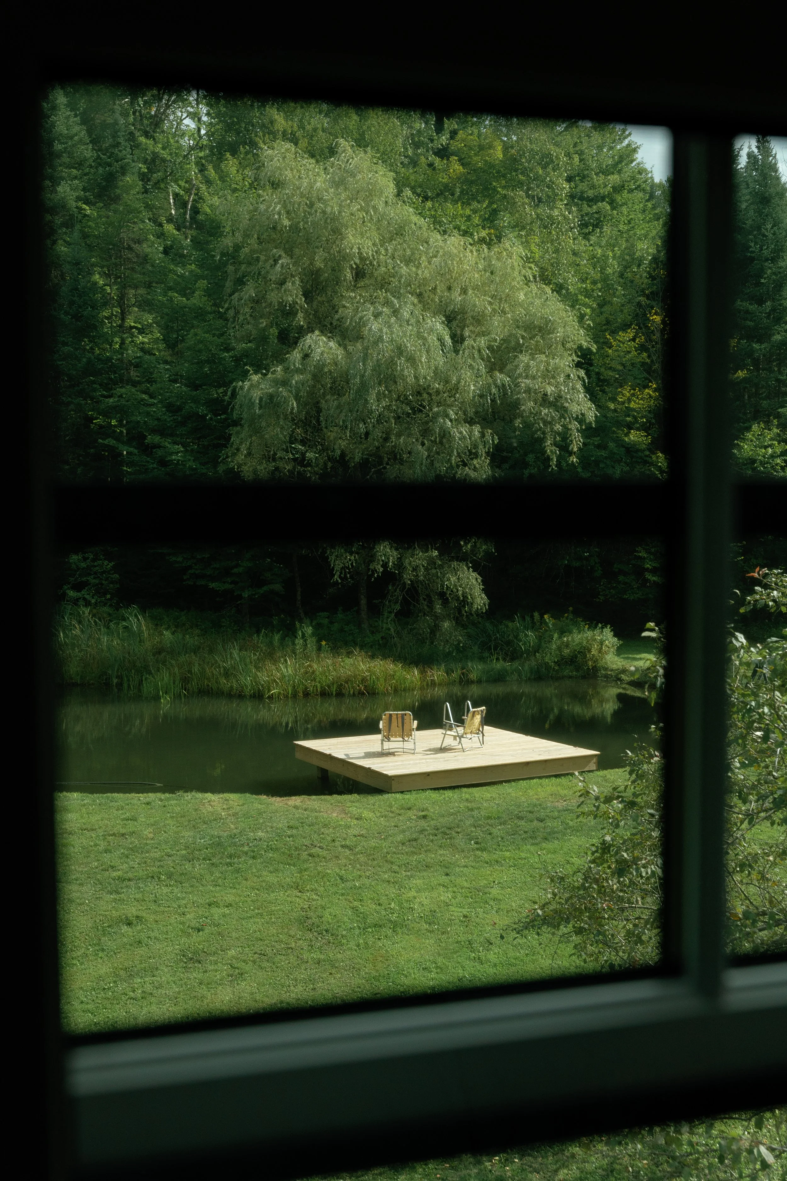 A view through a window showing a grassy yard with a floating dock and two chairs on it, on a calm pond, surrounded by dense trees and greenery.