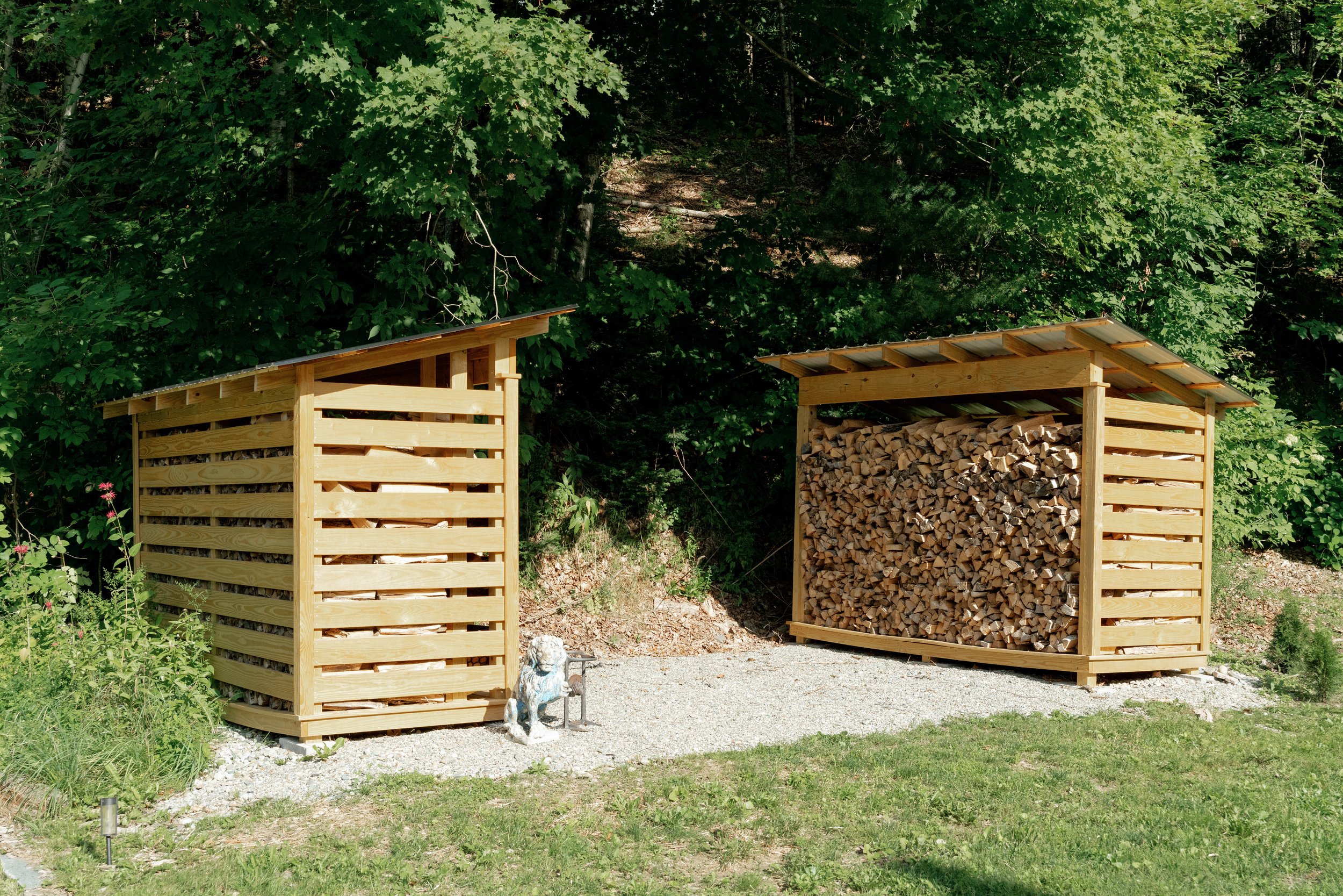 Two wooden wood storage sheds, one with a roof full of split firewood, set outdoors next to greenery and trees.