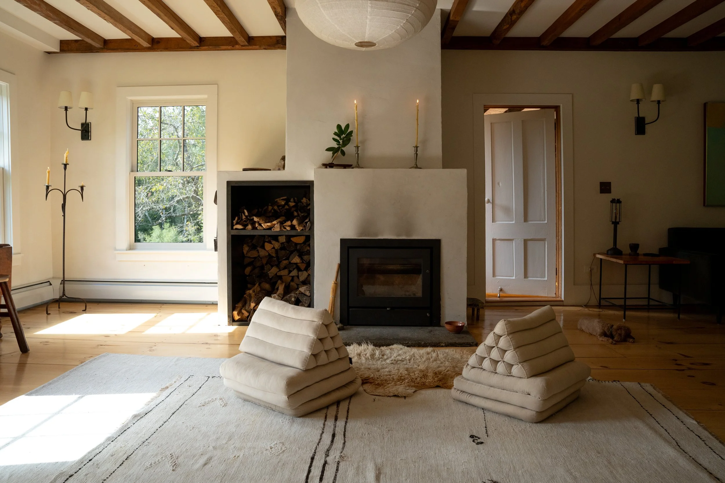 Living room with wooden beams, large window, white walls, wood fireplace with stacked firewood, candles, and cushions on the floor.