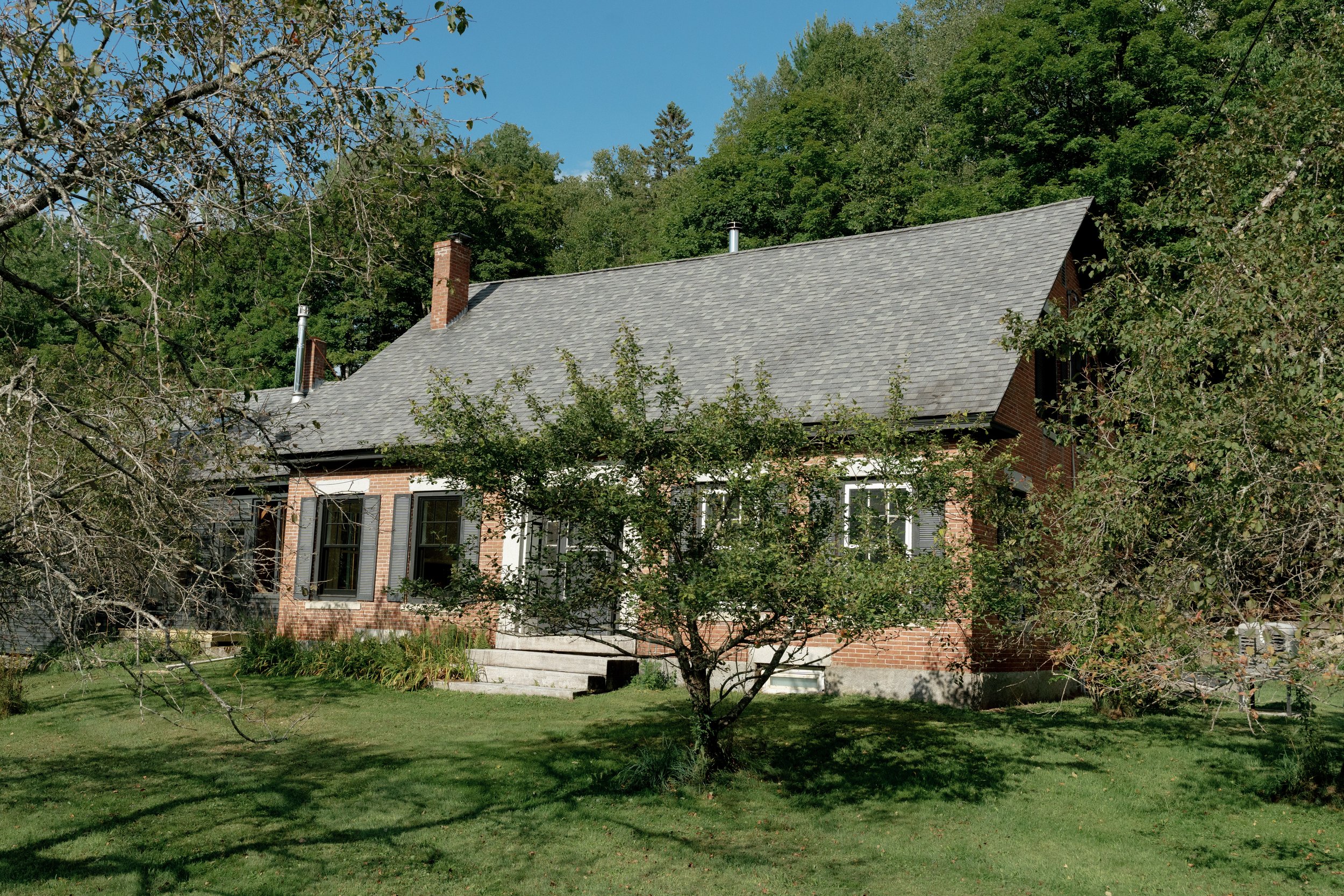 A brick house with a gray roof surrounded by trees and a green lawn, under a clear blue sky.