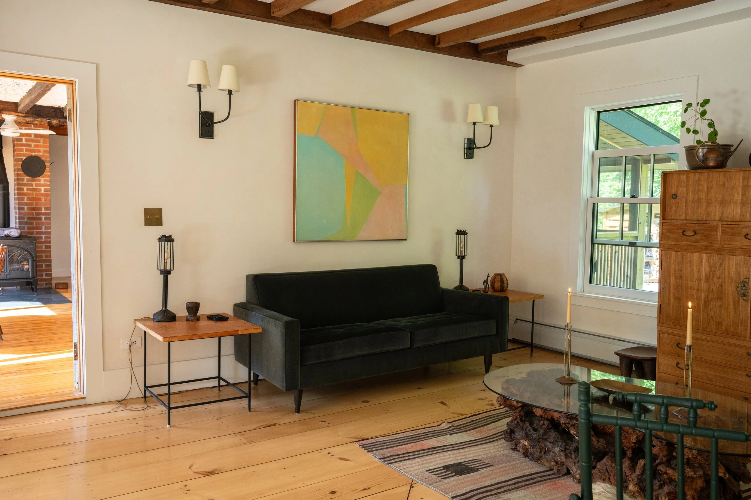 A cozy living room with wooden floors, white walls, and a wooden ceiling with exposed beams. There is a black sofa, a small wooden side table with decorative items, and a cabinet with pottery on top. The room has a window with a view of greenery outs