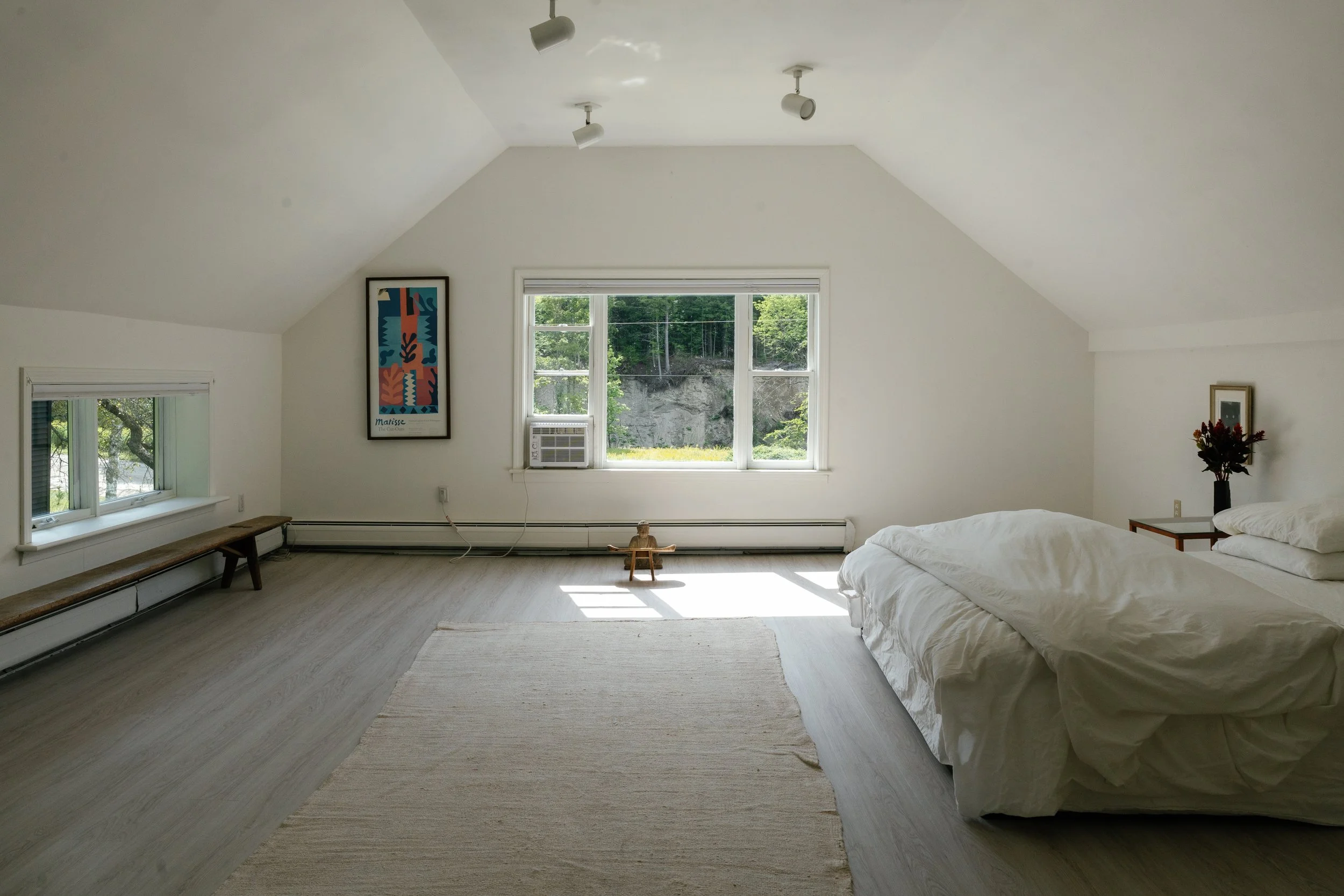 Minimalist bedroom with white walls, large window showing greenery, a bed with white bedding, a simple wooden side table with a vase of flowers, framed artwork on the wall, and ceiling spotlights.