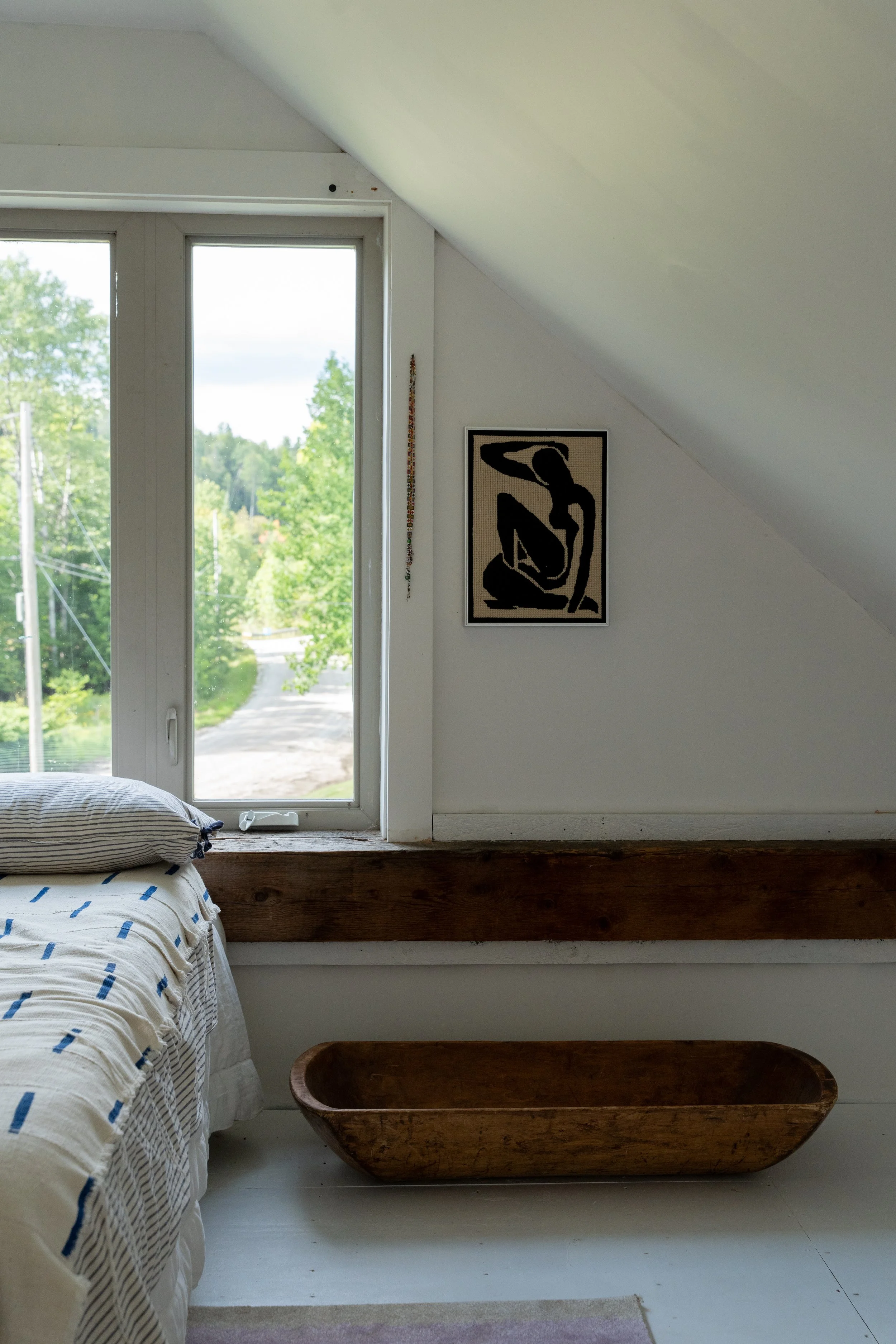 A cozy bedroom with a sloped ceiling, featuring a window opening to green trees and a cloudy sky. To the left, part of a bed with white bedding and blue stripes is visible. A framed black and beige artwork depicting a seated figure hangs on the wall.