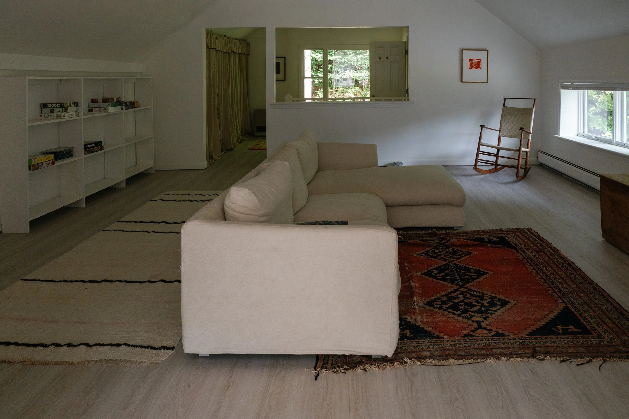 Living room with beige sofa, rocking chair near window, white bookshelf with books, and door opening to an outside view of greenery.