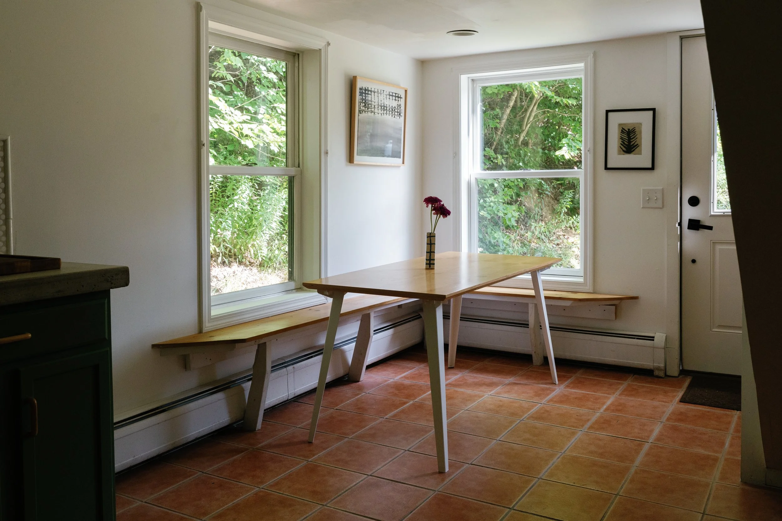 A bright kitchen with large windows showing outdoors greenery. There is a wooden dining table with a small vase of purple flowers, a corner of green cabinetry, and framed art on the walls. The floor has reddish-brown tiles.