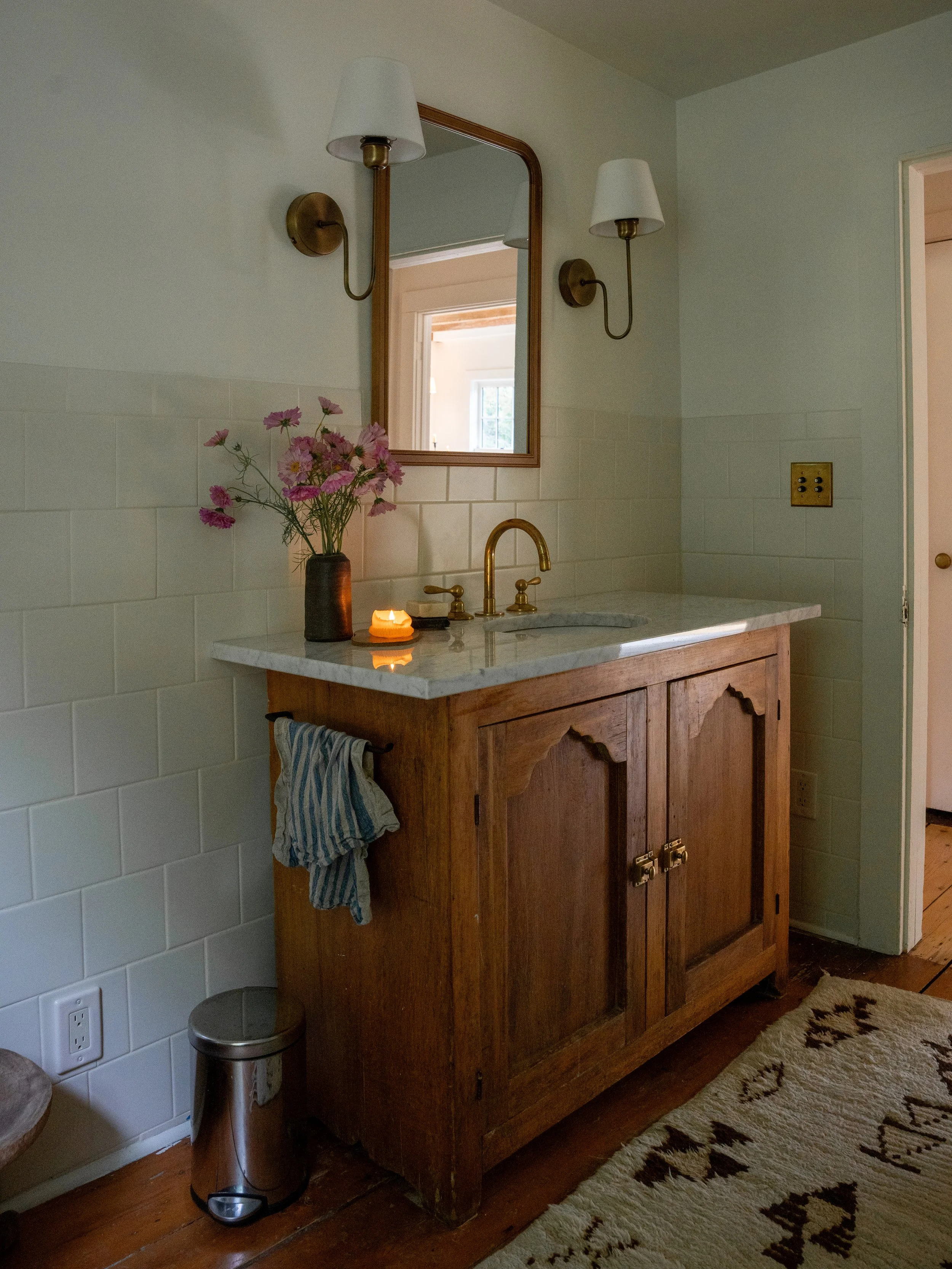 A bathroom vanity with a wooden cabinet, a marble countertop, a brass faucet, a mirror, and two wall-mounted lamps with white shades. There's a bouquet of pink flowers in a dark vase, a lit candle, a towel hanging on the cabinet's side, and a beige r