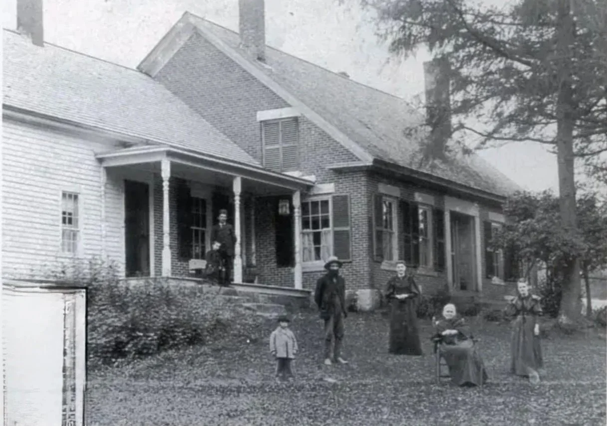 Vintage black and white photo of a large house with a group of six people, including three children and three adults, standing on the front yard. The house has a gabled roof, multiple windows, and a porch with columns.