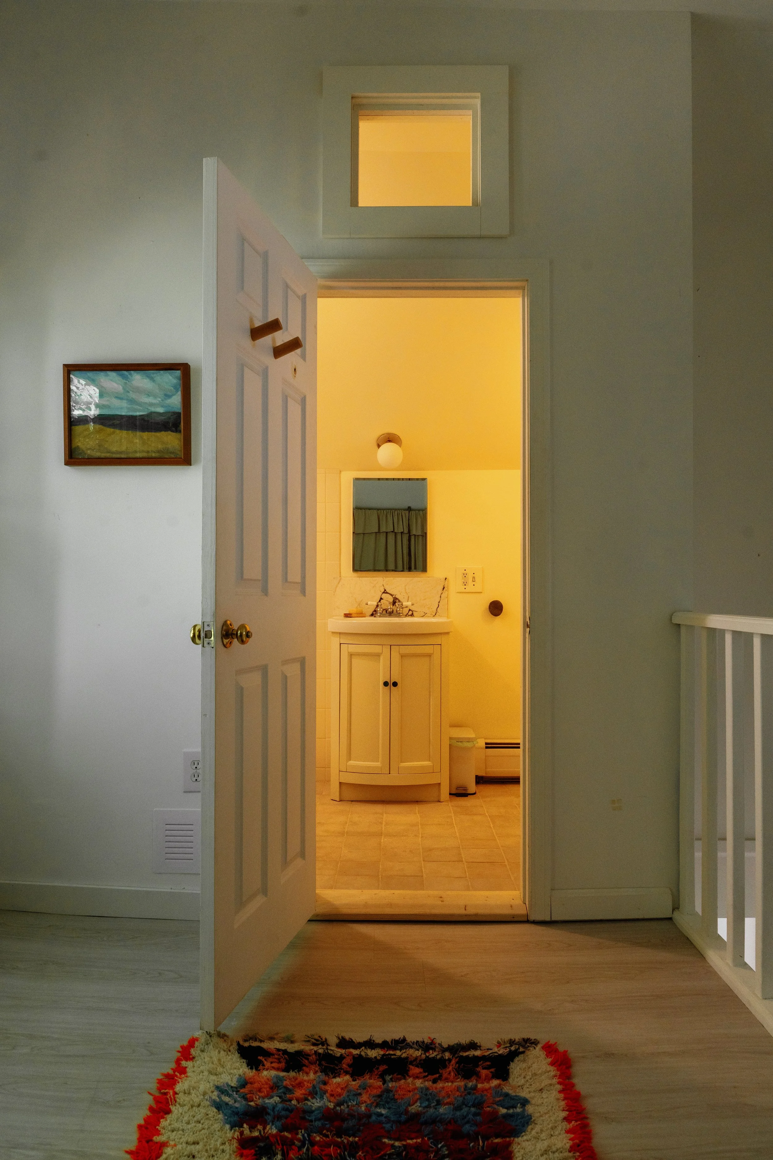 View from a living area looking into a bathroom through an open door. The bathroom has a small vanity with a mirror, a sink, and yellow walls. The hallway and living area have wooden flooring, with a colorful rug in the foreground.