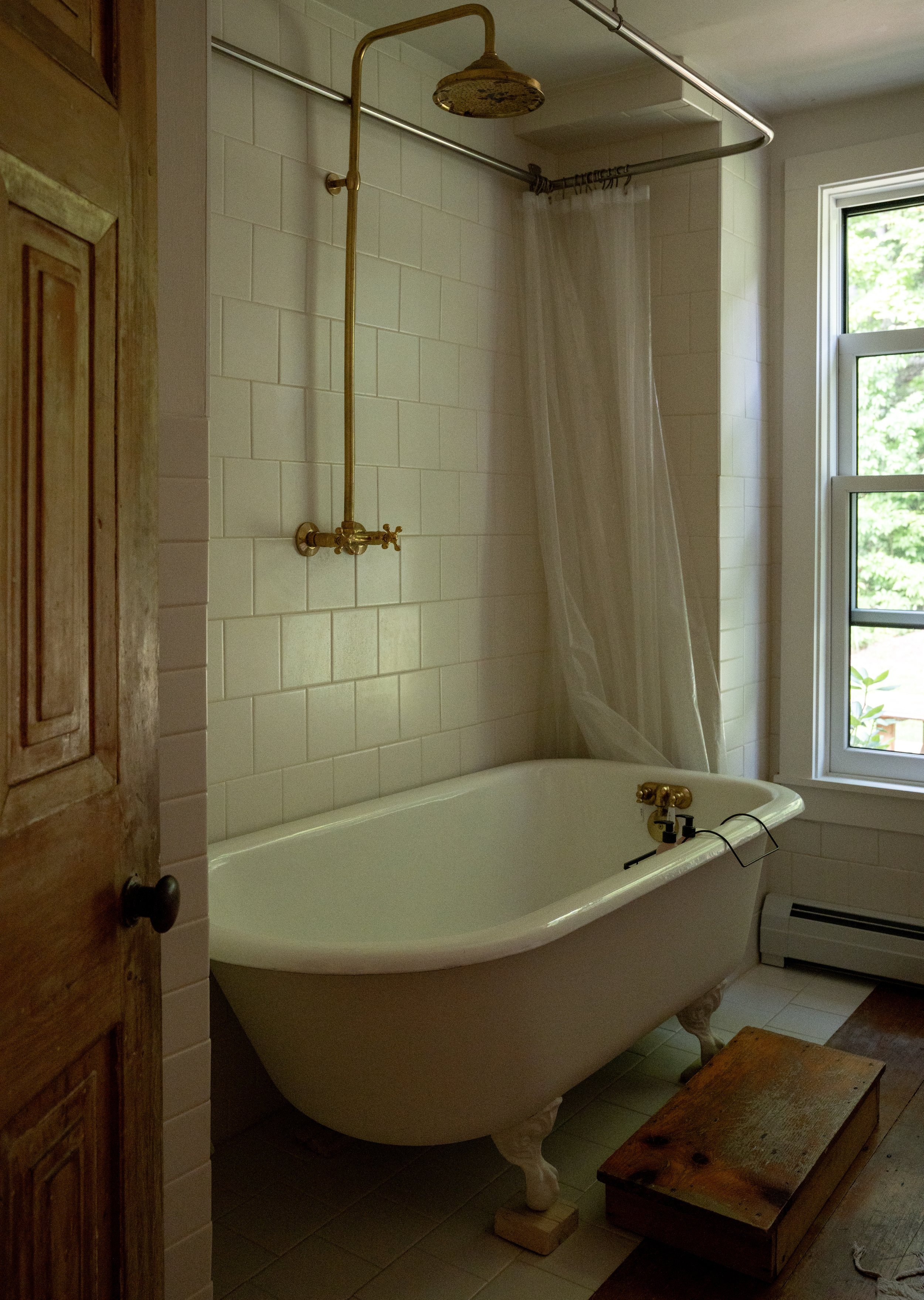 A vintage clawfoot bathtub with brass fixtures in a bathroom, with a window showing greenery outside, a wooden stool nearby, and a framed mirror on the wall.