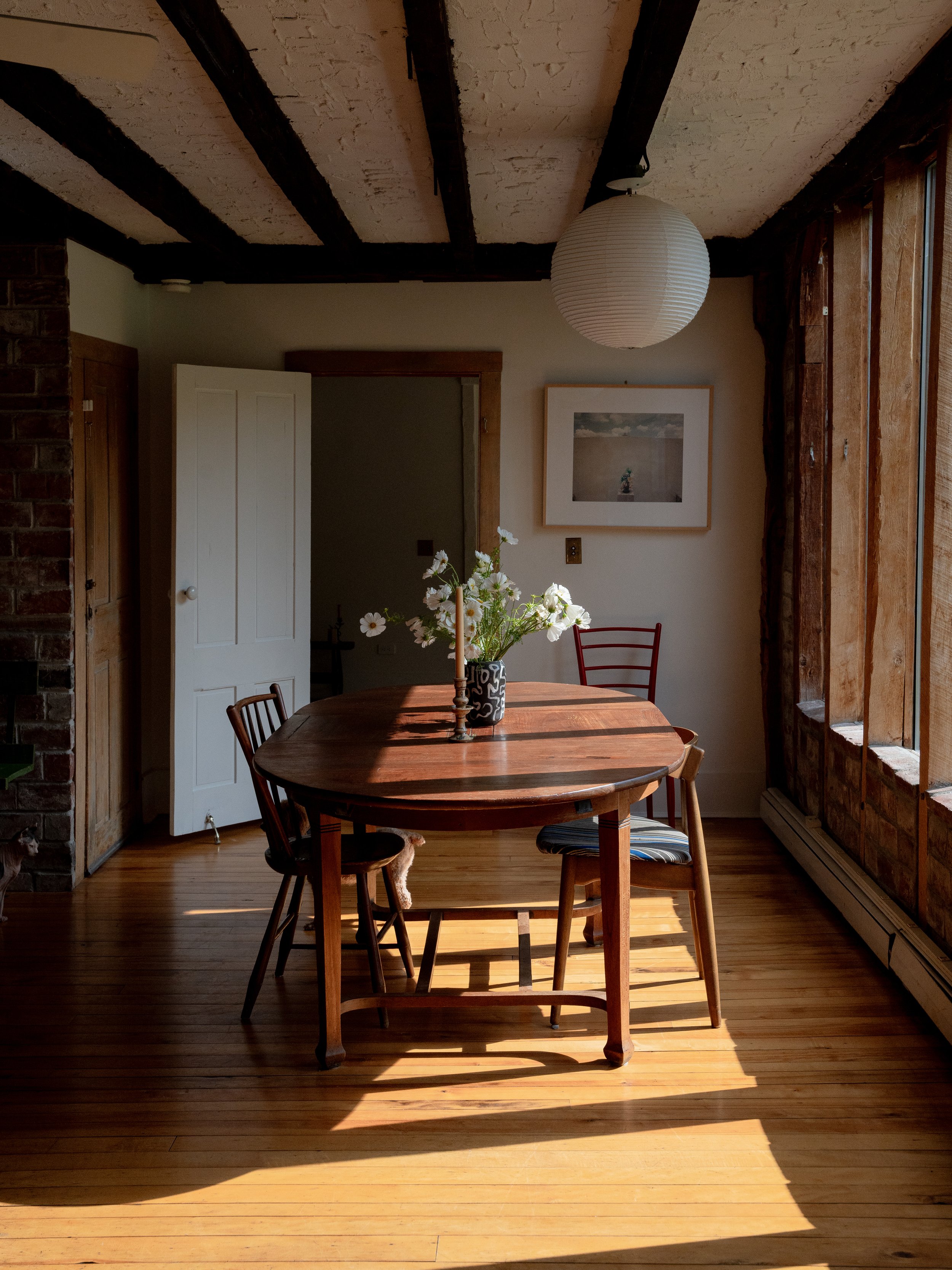 A cozy dining area with a wooden table, four wooden chairs, a bouquet of white flowers in a black and white vase, and a white paper lantern hanging from the ceiling. Sunlight streams through large windows on the right, casting shadows on the wooden f