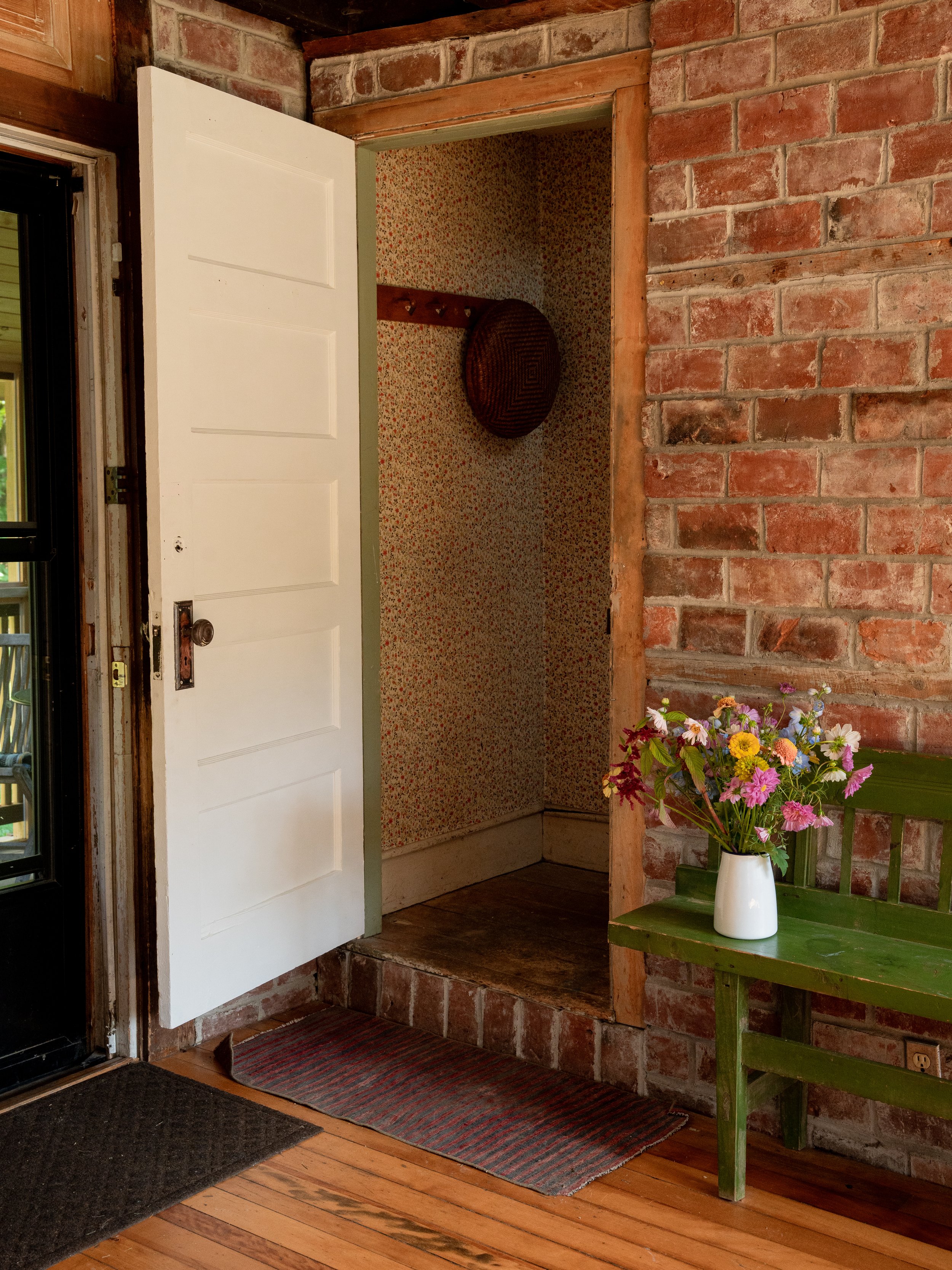 View of a front porch area with a brick wall, a green wooden side table with a vase of colorful flowers, a white door propped open revealing a small closet with floral wallpaper and hooks. A doormat and hardwood floor are visible.