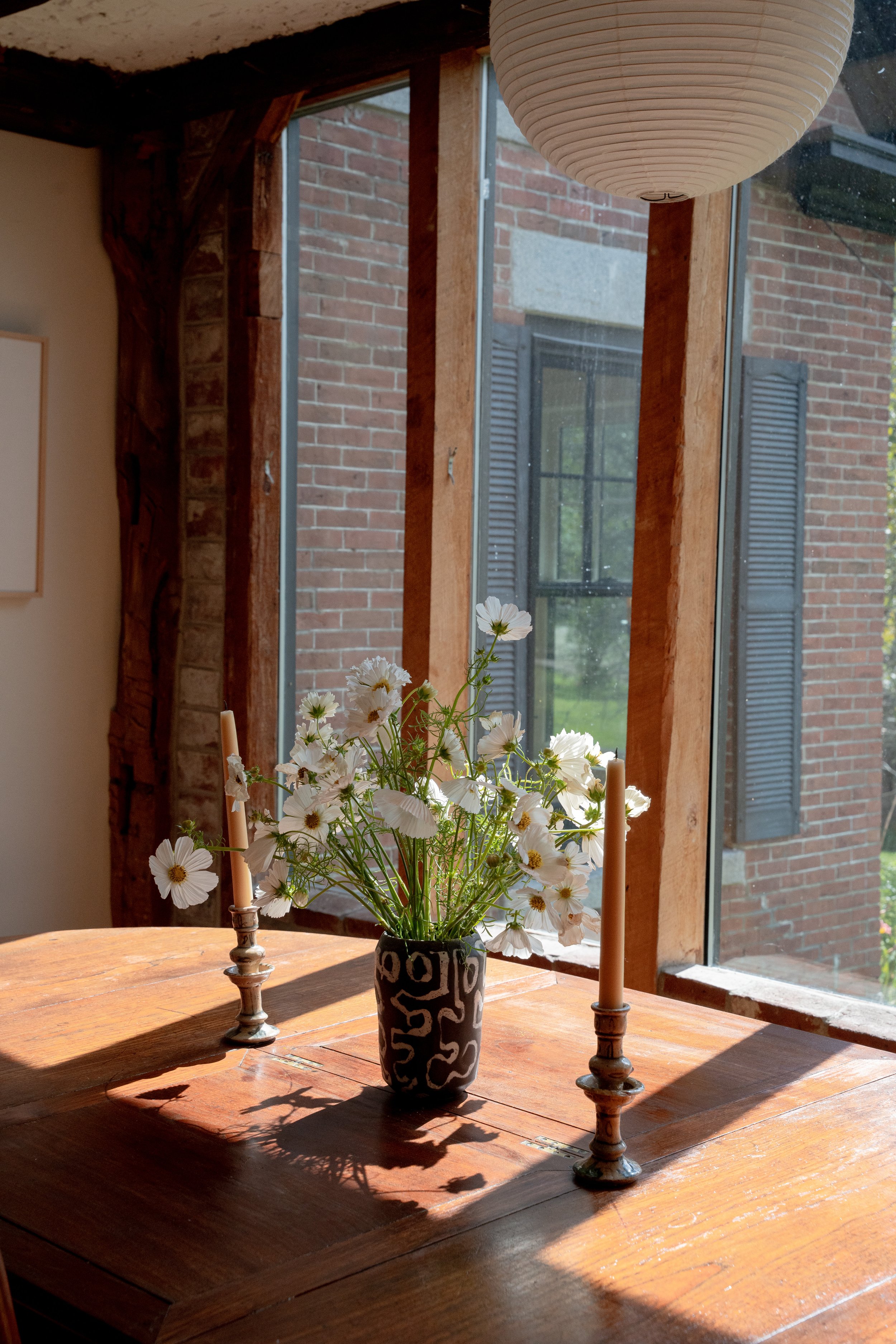A wooden dining table with a black and white vase containing white flowers and two tall candlesticks with candles, placed in front of large windows with brick buildings outside, in natural daylight.