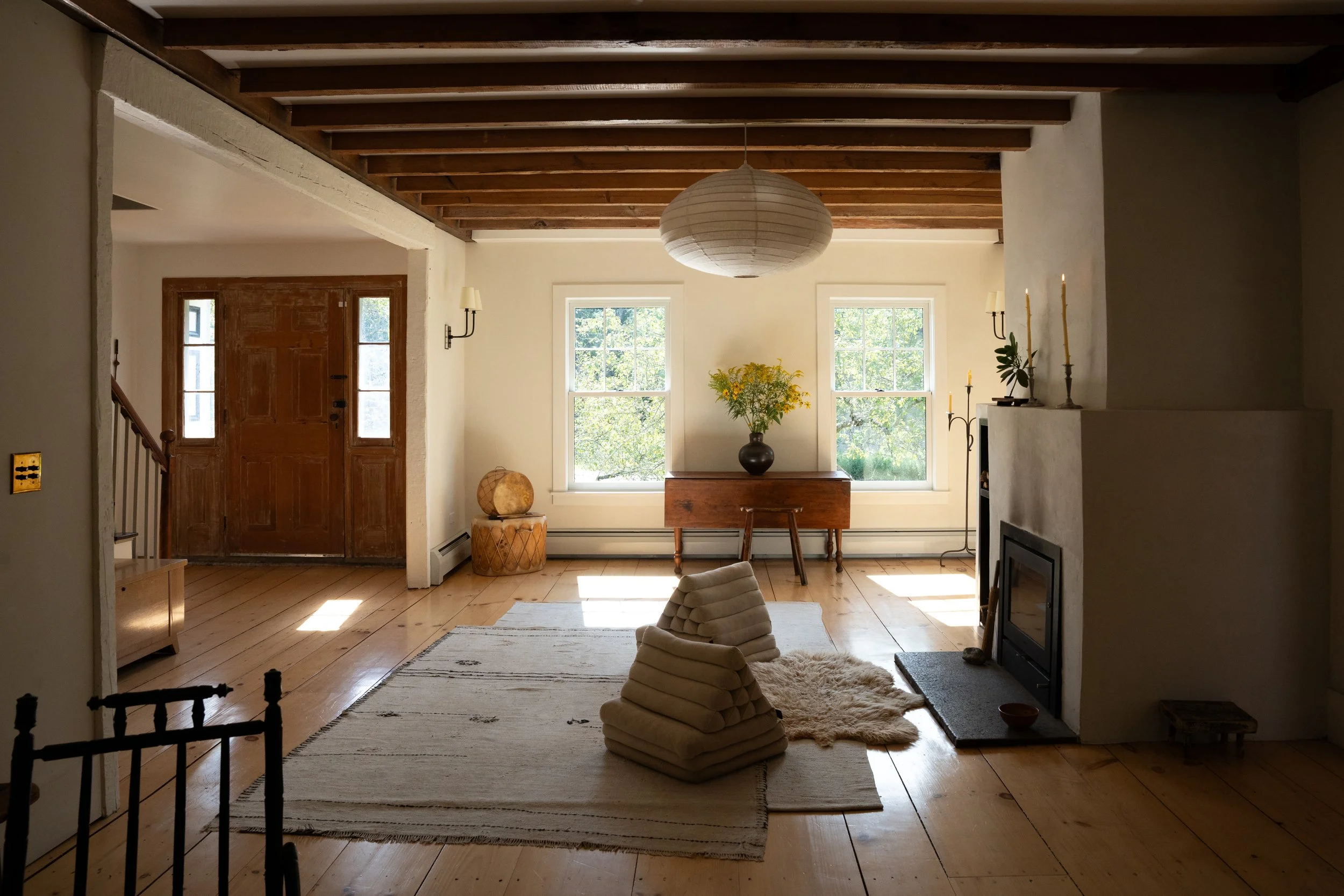 Living room with wooden floor, white walls, large windows, a wooden front door, houseplants, a table, chairs, a cozy carpet, a wrapped fabric decor, a hanging paper lantern, and candles on a mantle.